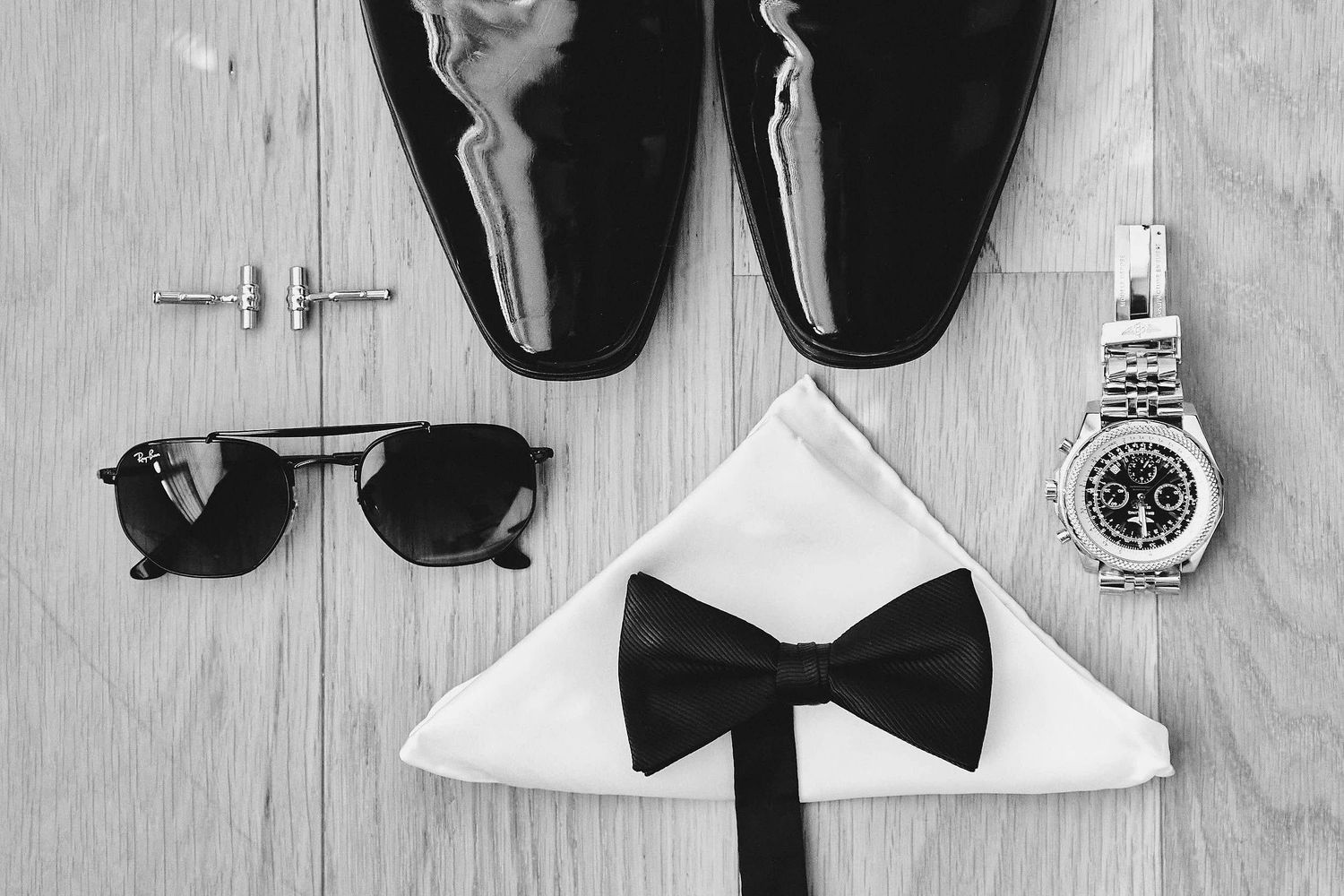 Formal black and white accessories including dress shoes, sunglasses, bow tie, and watch arranged on wooden surface.