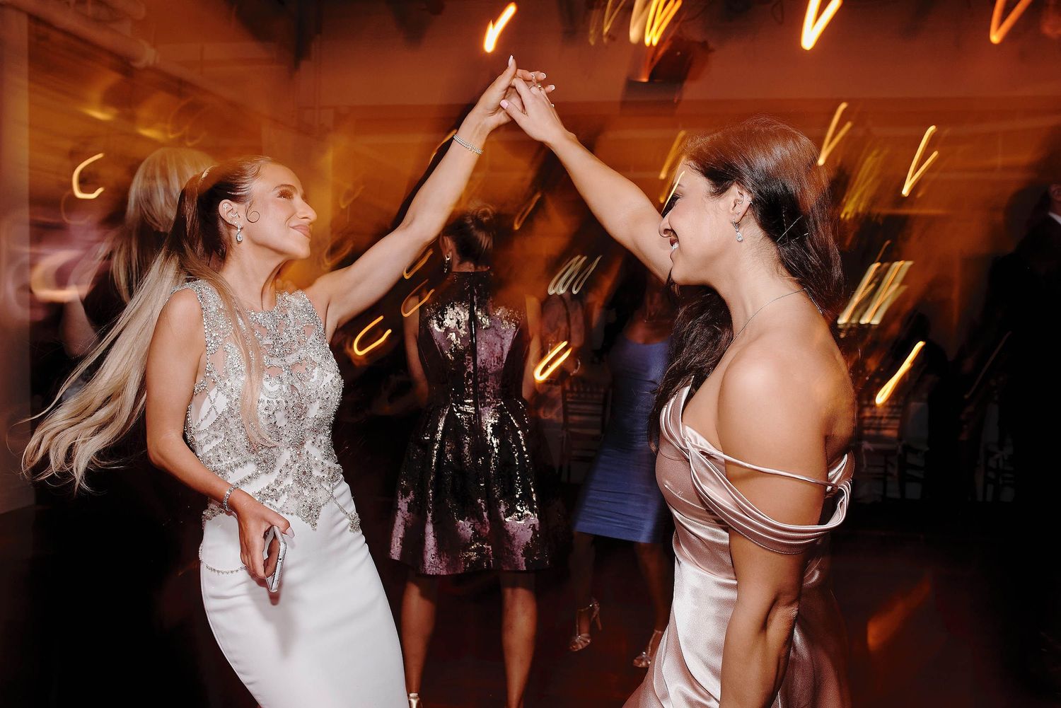 Two guests in elegant white gowns dance together with raised arms on a festive dance floor with blurred lights in background.