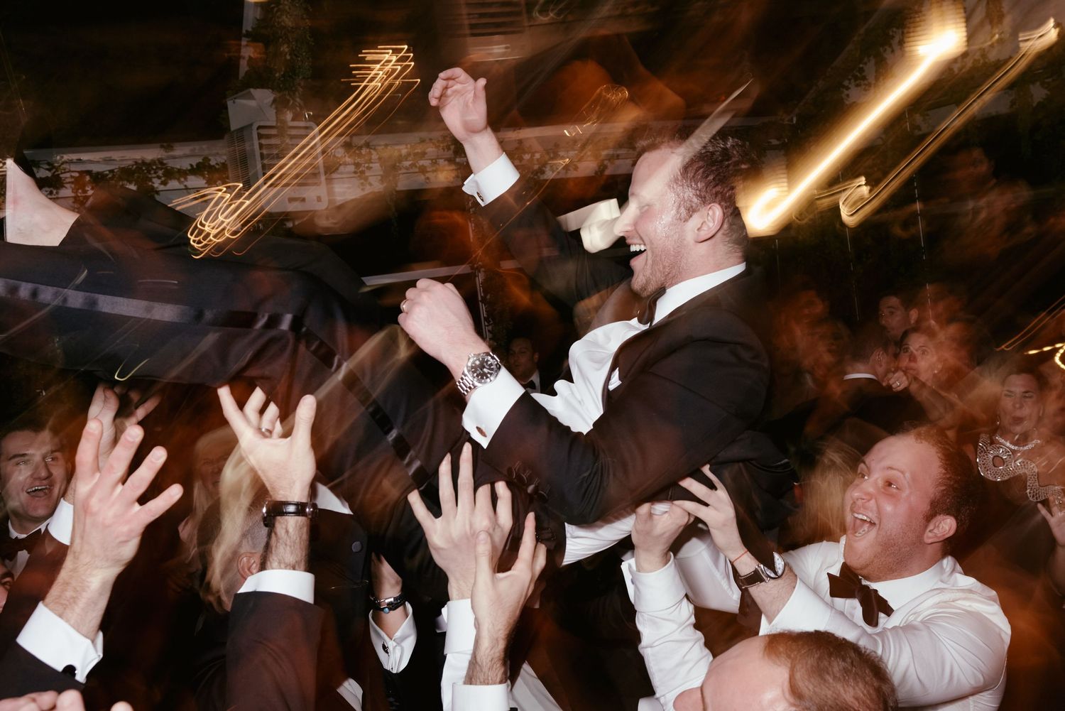 A crowd of wedding guests in formalwear raise their hands in celebration at a lively reception party.