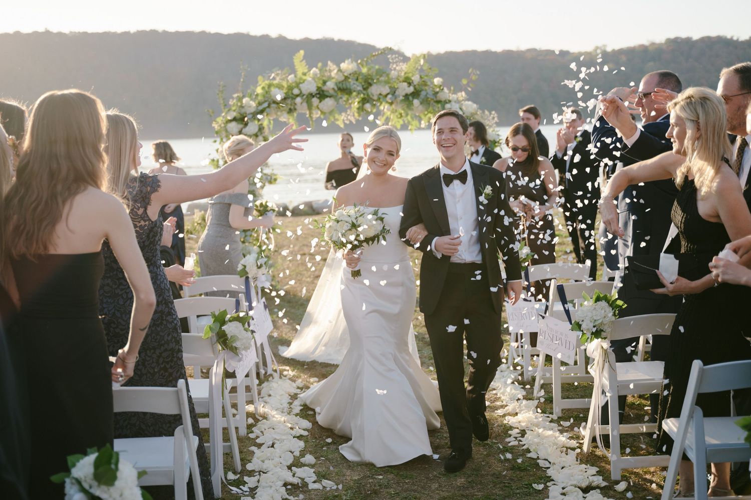 Wedding couple walks down aisle while guests throw confetti during sunset outdoor ceremony.