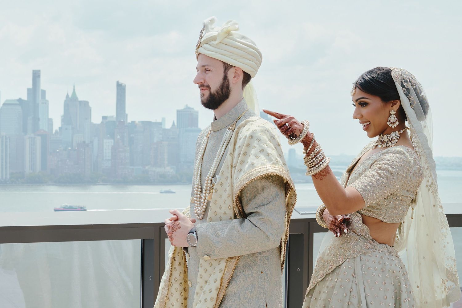 A couple in traditional South Asian wedding attire share a moment on a rooftop with city skyline in background.