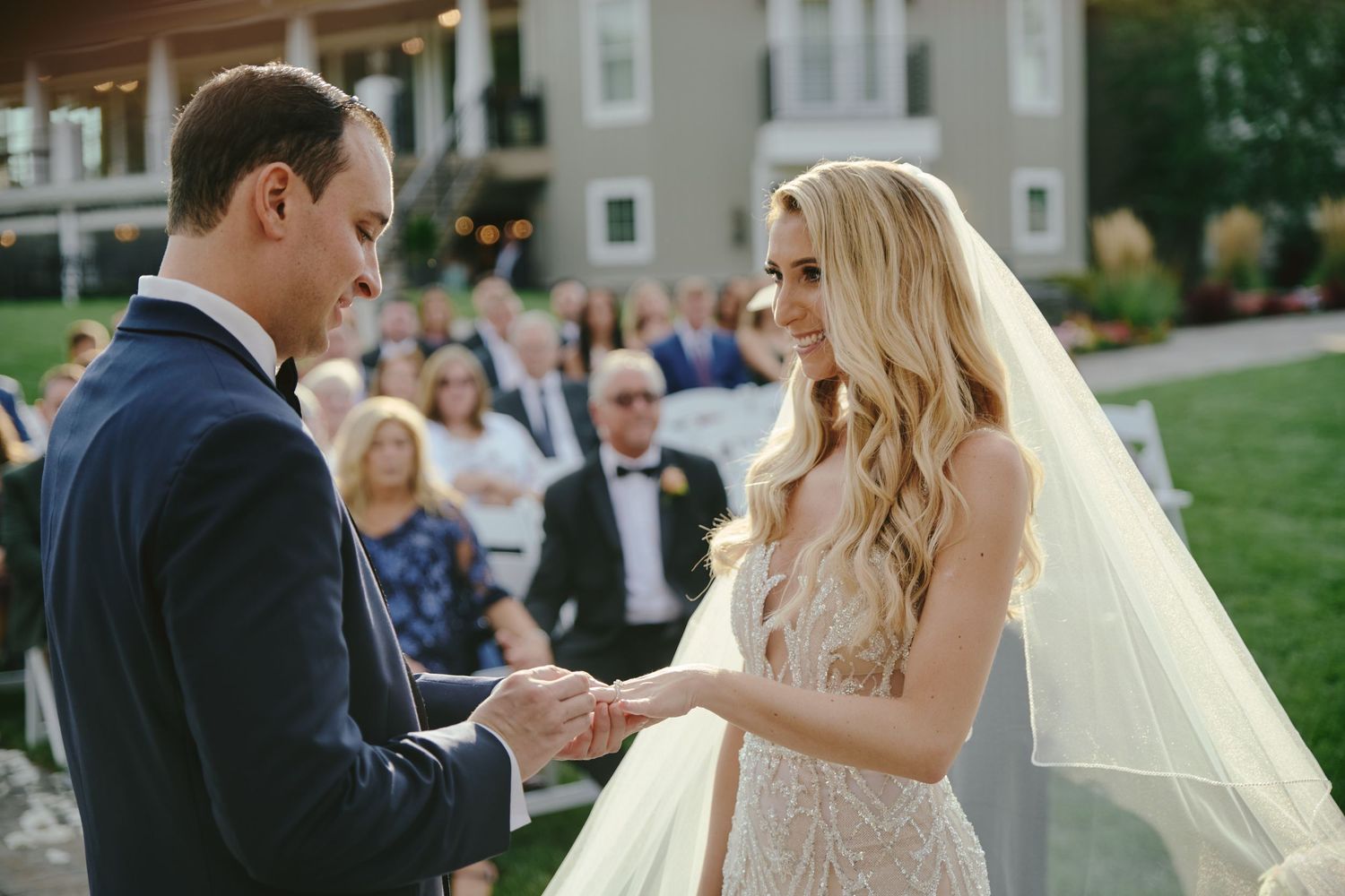 A bride and groom exchange rings during an outdoor wedding ceremony while guests look on from their seats.