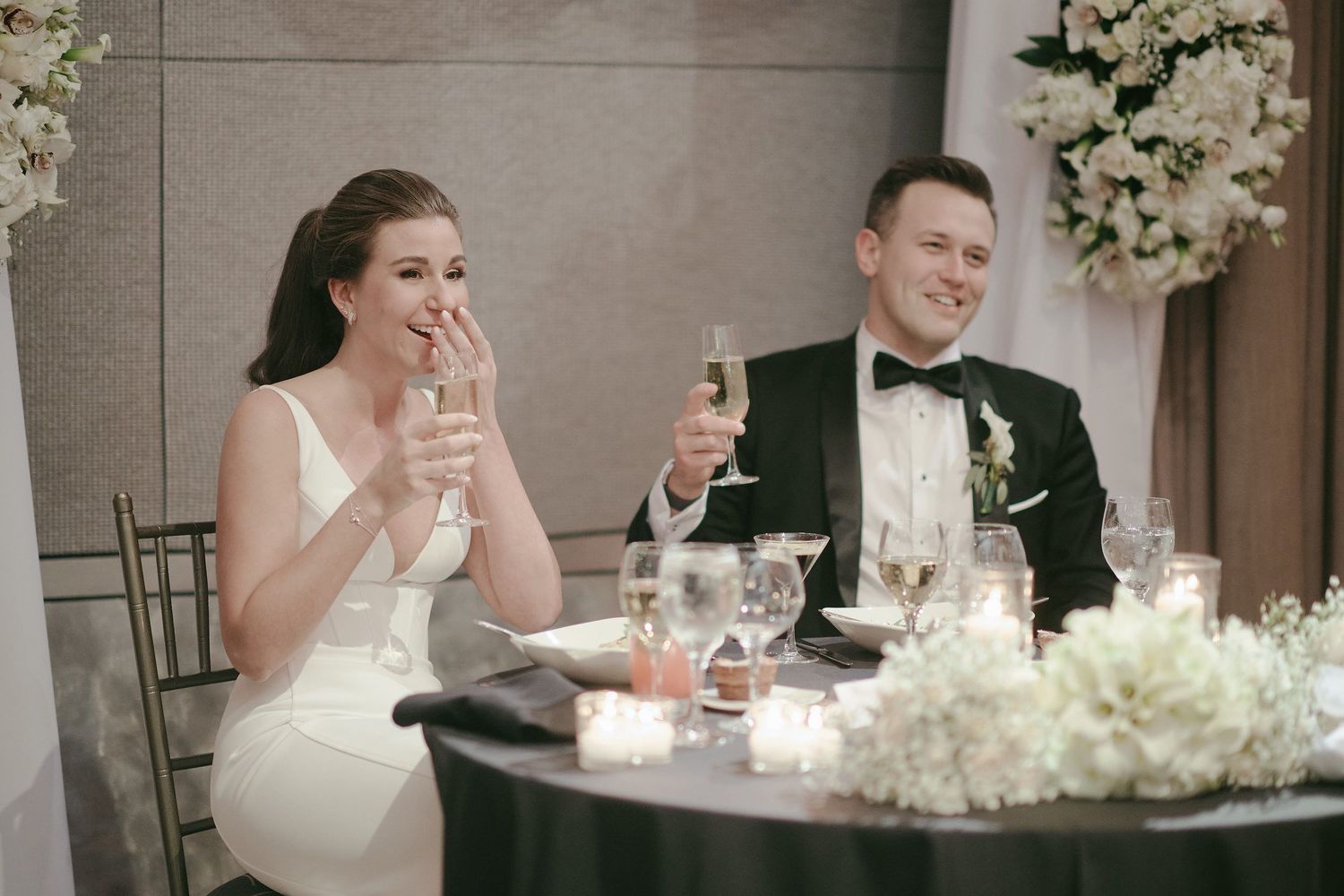 Newlyweds share a laugh while raising champagne glasses at their elegantly decorated reception table.