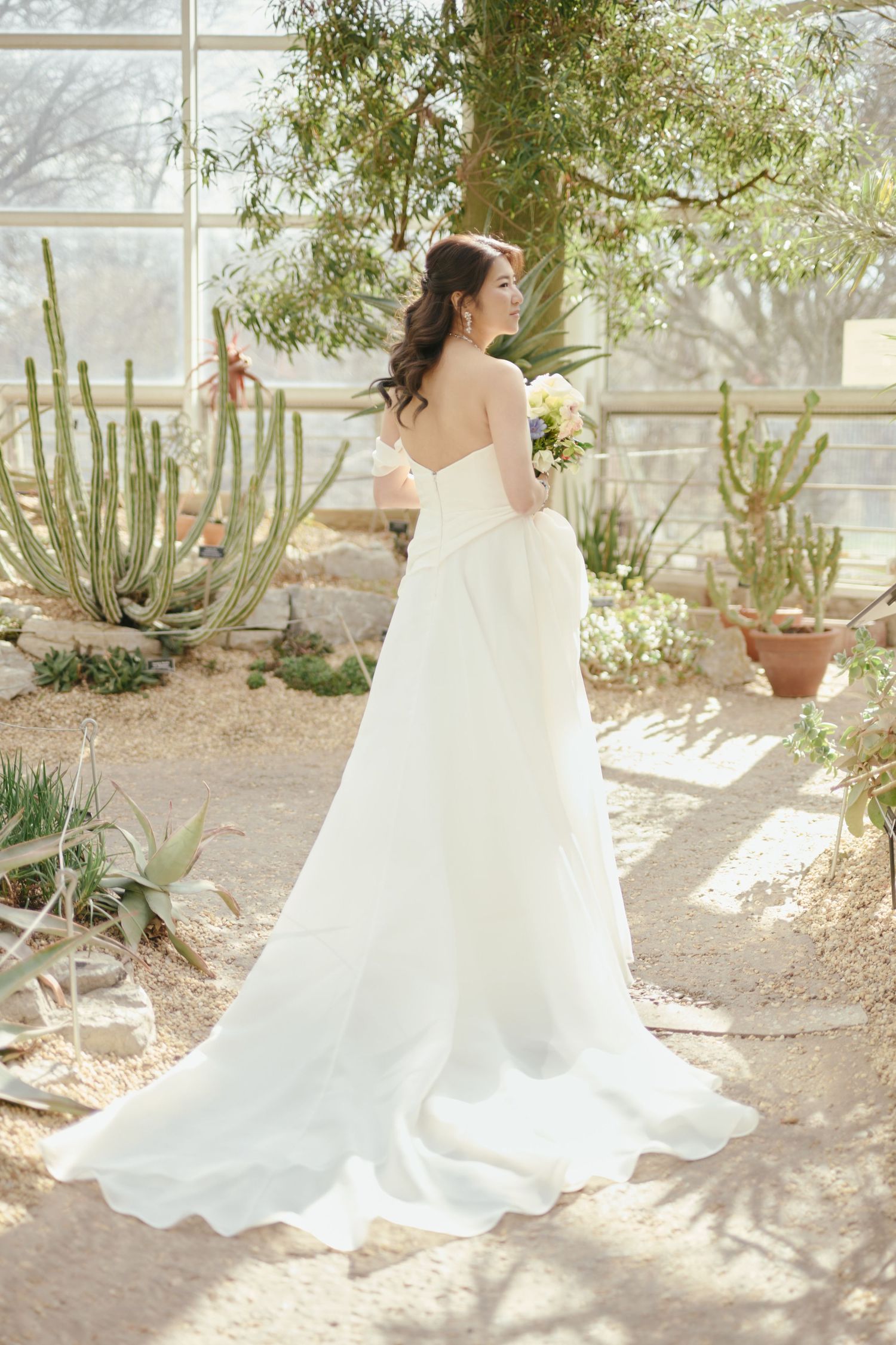 Bride in strapless white gown with train holds bouquet in sunlit greenhouse surrounded by cacti and desert plants.