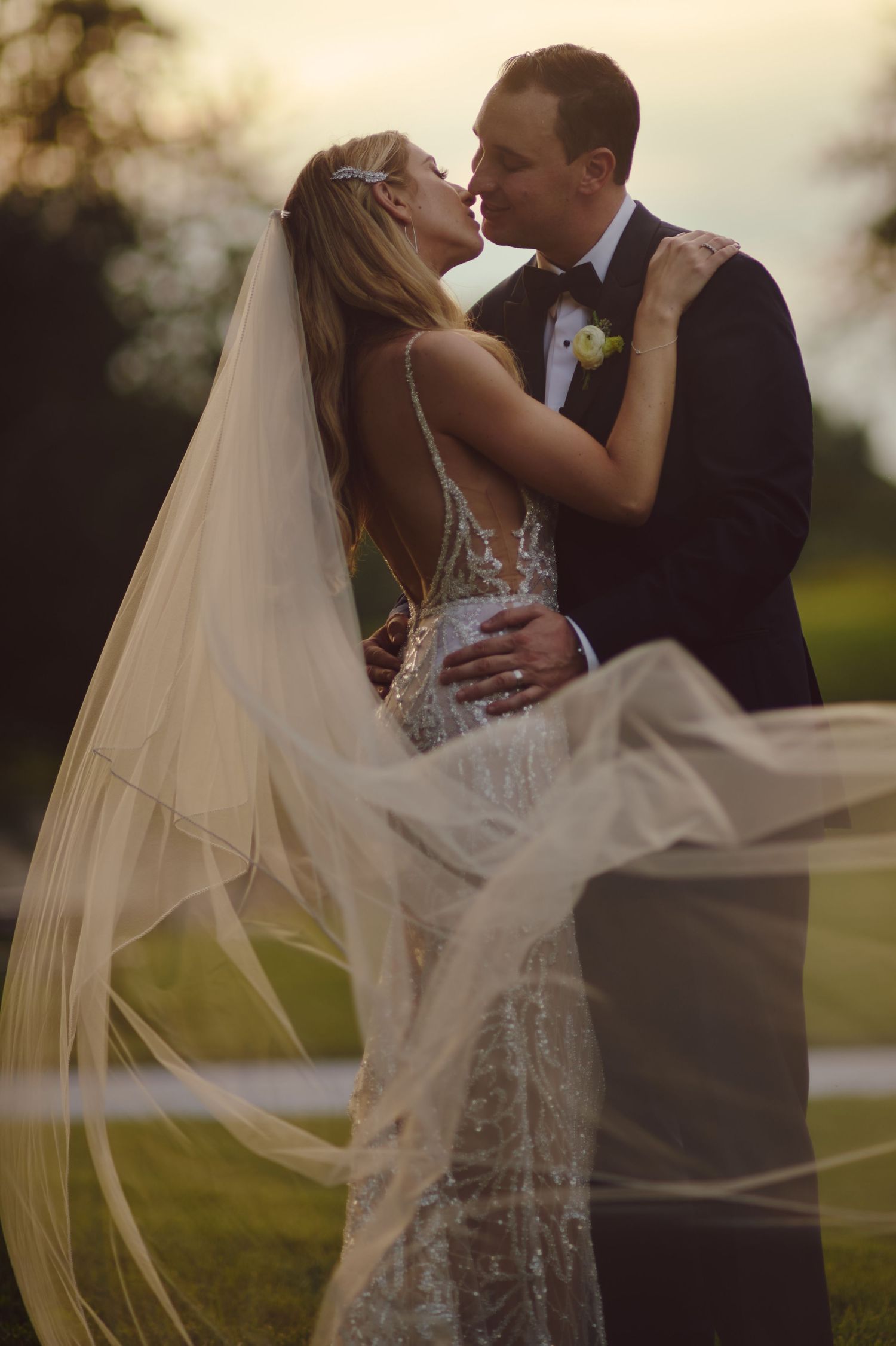 A romantic newlywed couple shares an intimate kiss at sunset while the bride's long veil flows in the breeze.