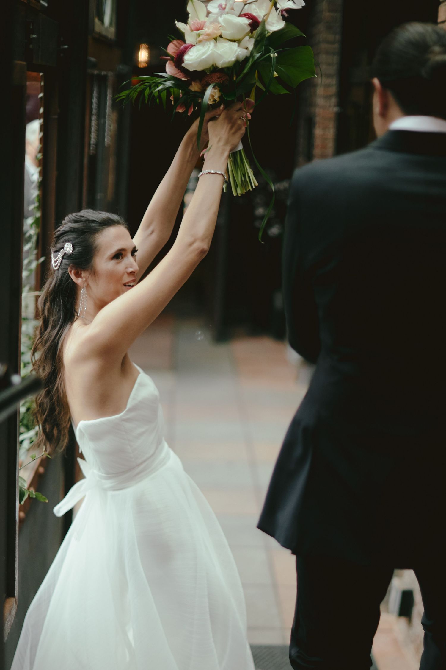 A bride in a white wedding dress raises her bouquet of pink and white flowers triumphantly during the celebration.