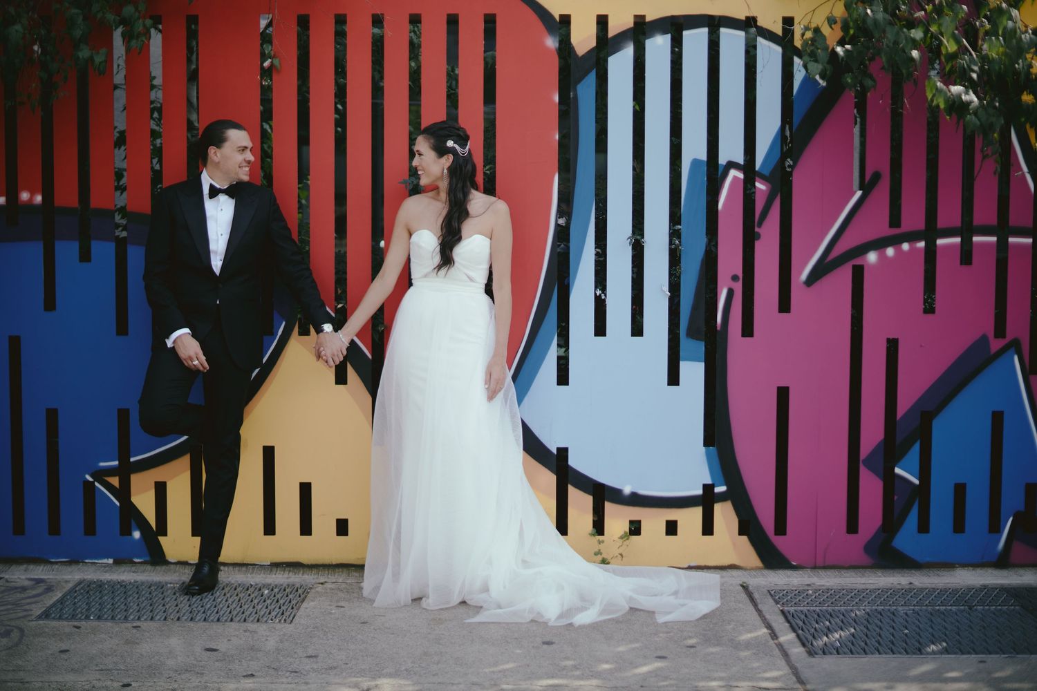 A couple poses together against a vibrant colorful mural wall, with the bride in a flowing white gown.