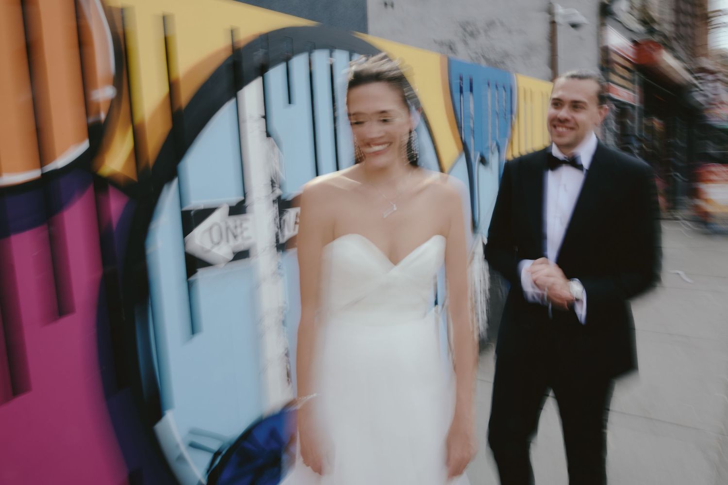 A joyful bride in a strapless white gown and person in black tuxedo stand in front of colorful graffiti wall.