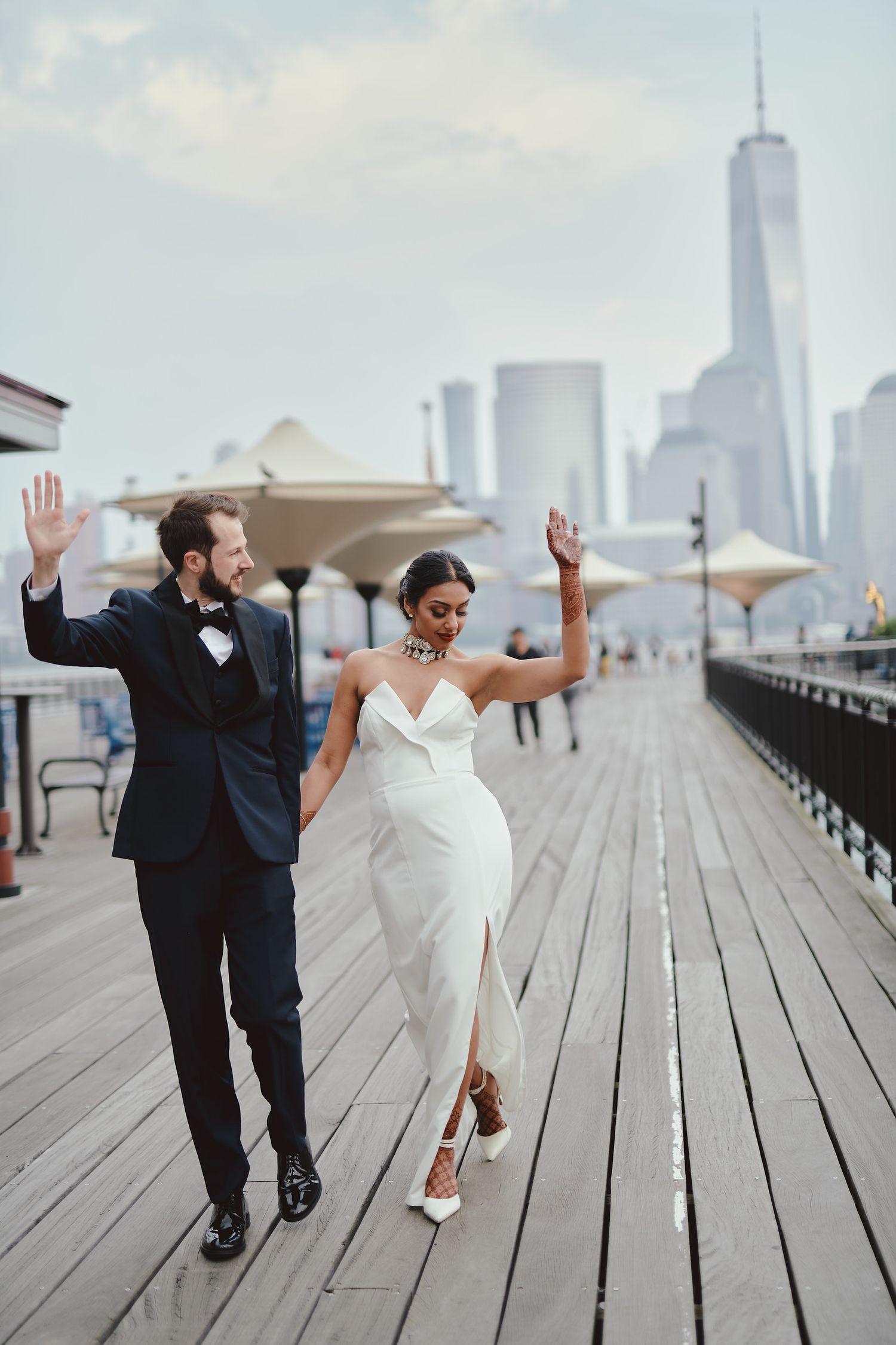 Newlywed couple joyfully dancing on a waterfront boardwalk with the Freedom Tower and NYC skyline in the background.
