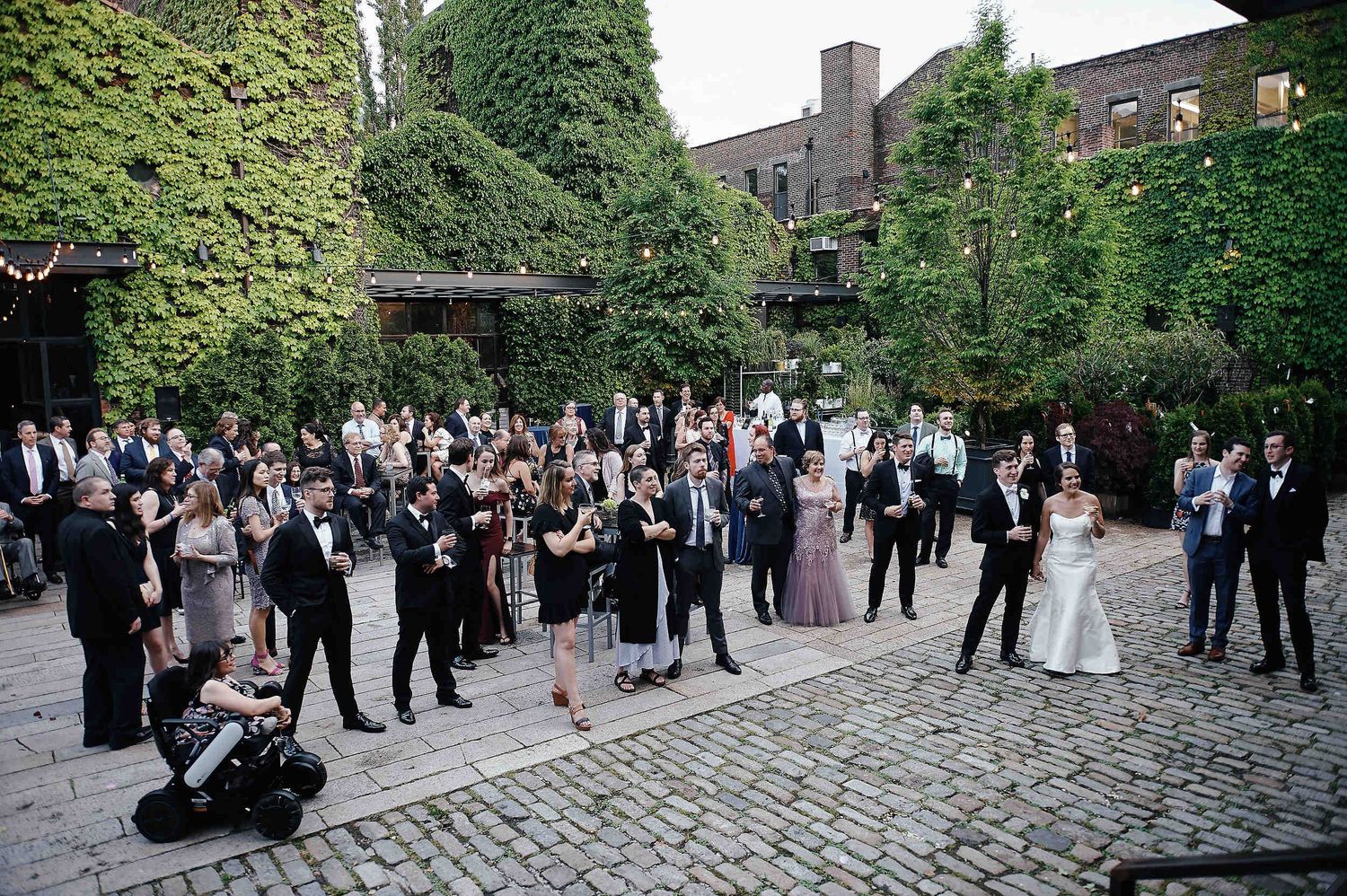 Large wedding party gathering in an ivy-covered courtyard with cobblestone ground during an outdoor celebration.