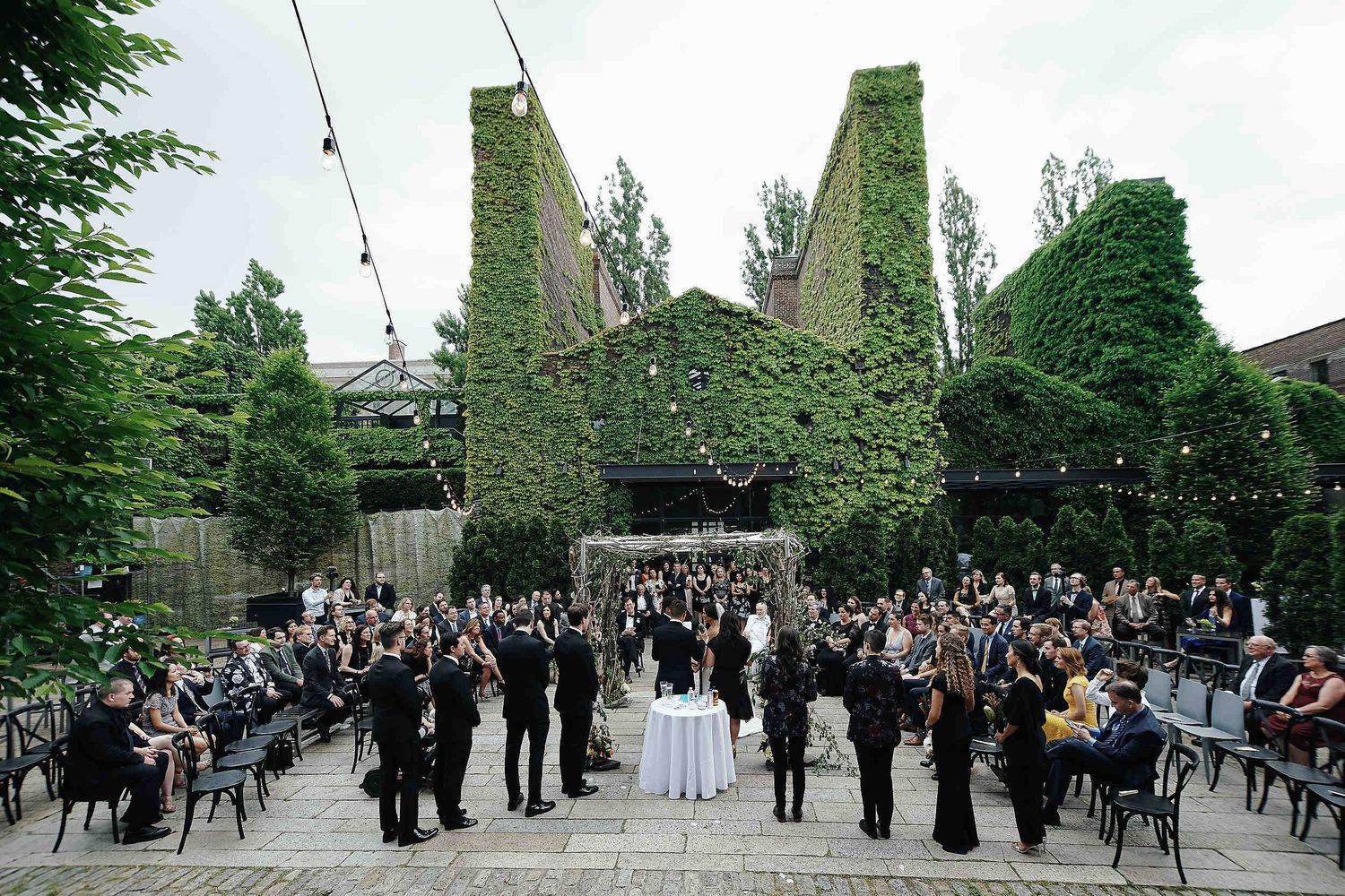 An outdoor wedding ceremony setup with ivy-covered pillars and guests seated in chairs on a paved courtyard.