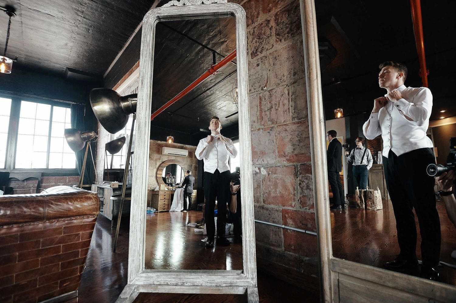 Person in white button-up shirt adjusts clothing in vintage mirror while others prepare in rustic industrial space.