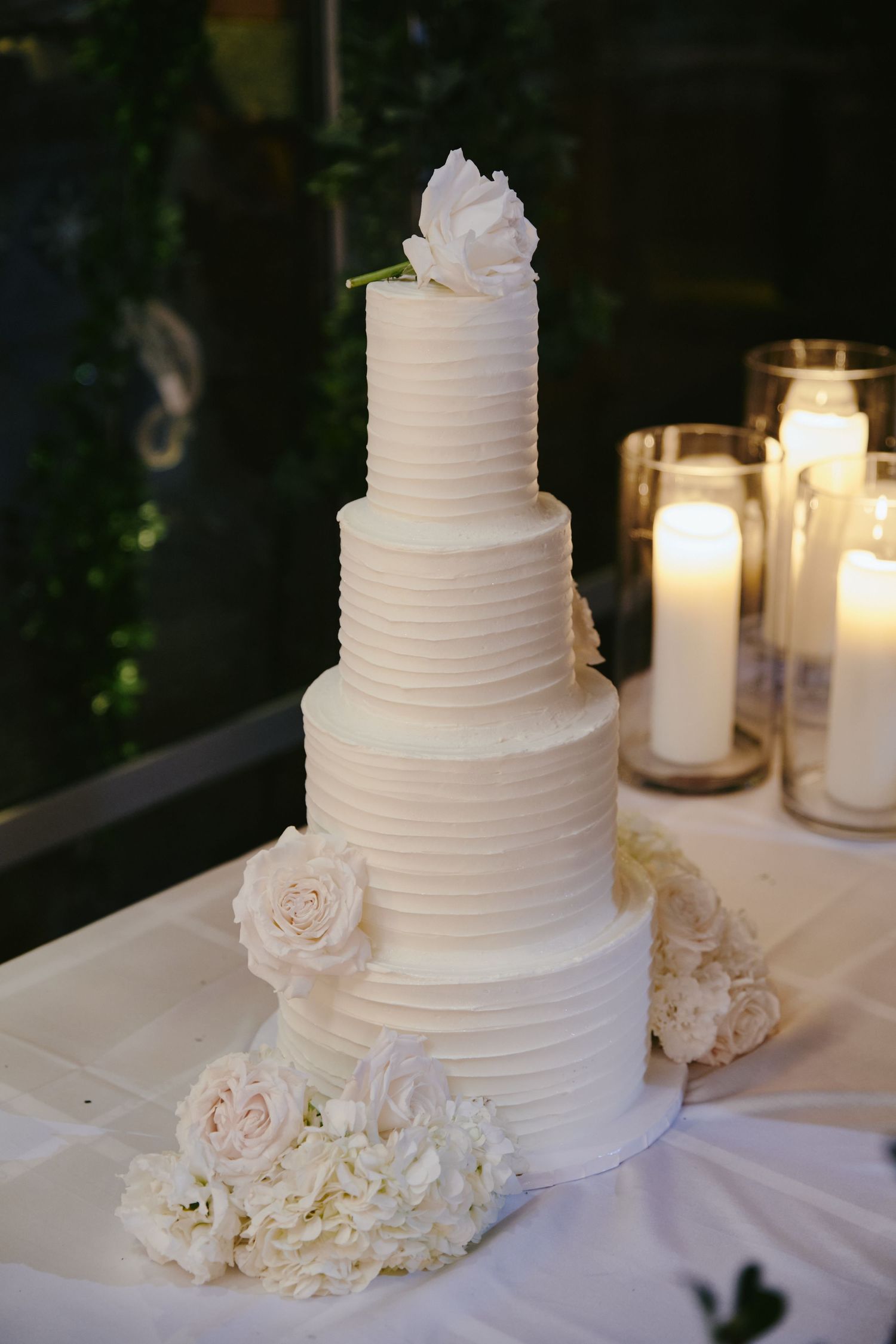 Elegant four-tier white wedding cake with textured frosting and fresh white roses, displayed next to glowing candles.