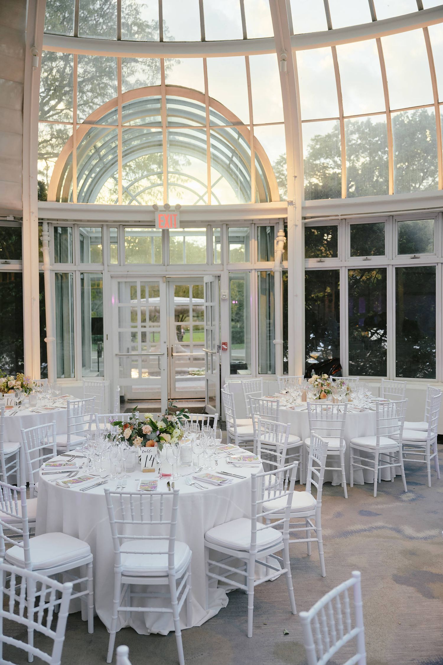 Elegant wedding reception setup in a glass conservatory with white chiavari chairs and round tables under arched windows.