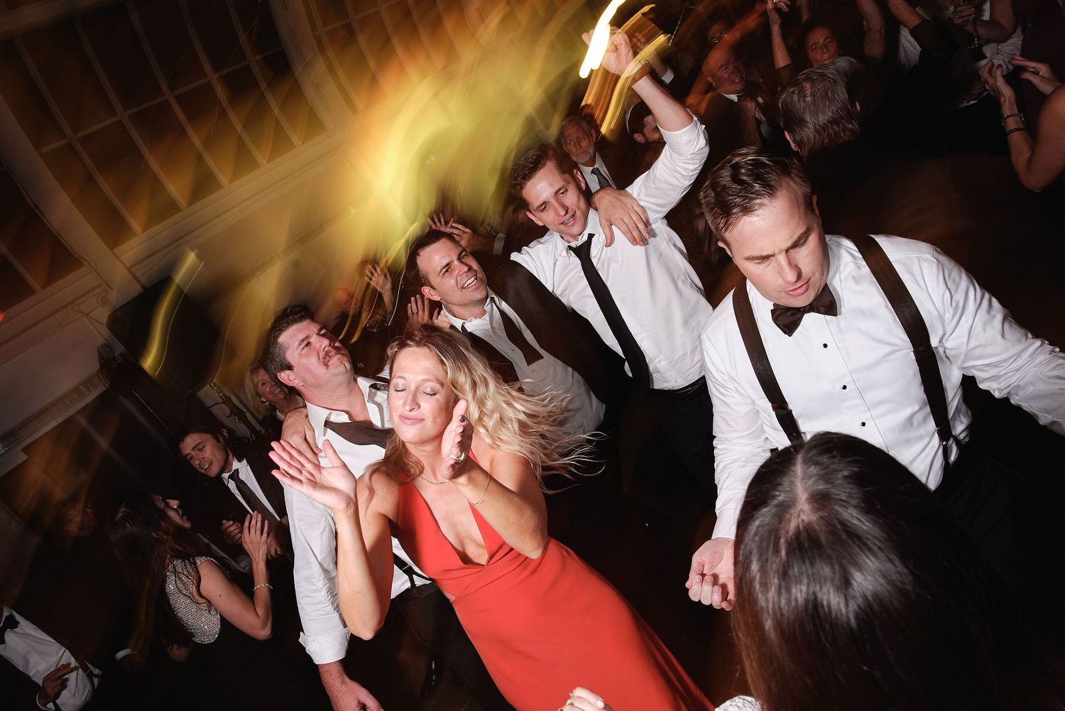 Wedding reception party guests dancing enthusiastically on a dance floor with dramatic lighting in background.