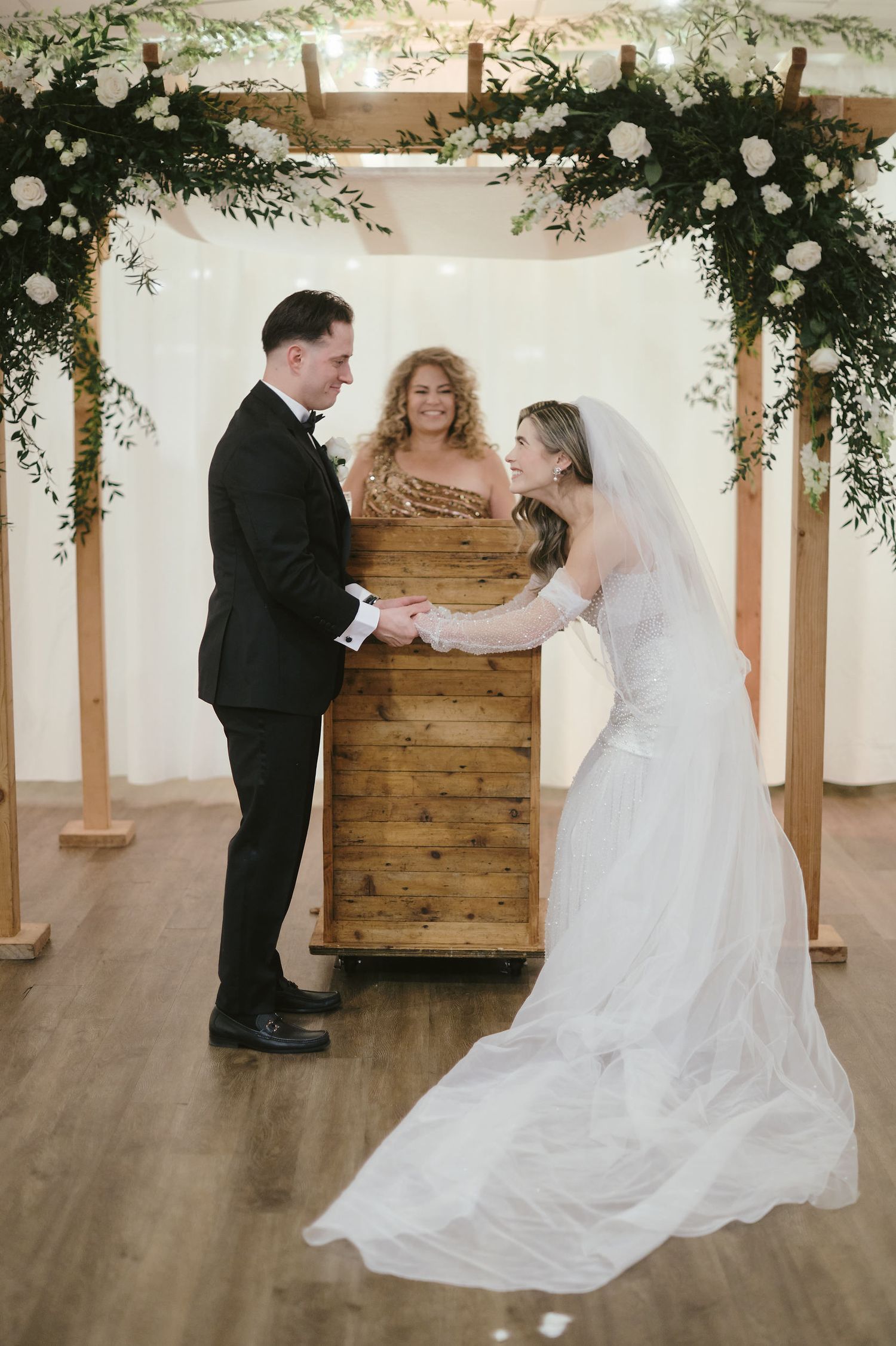 A wedding ceremony takes place under a floral arch with greenery and white roses against a wooden backdrop.