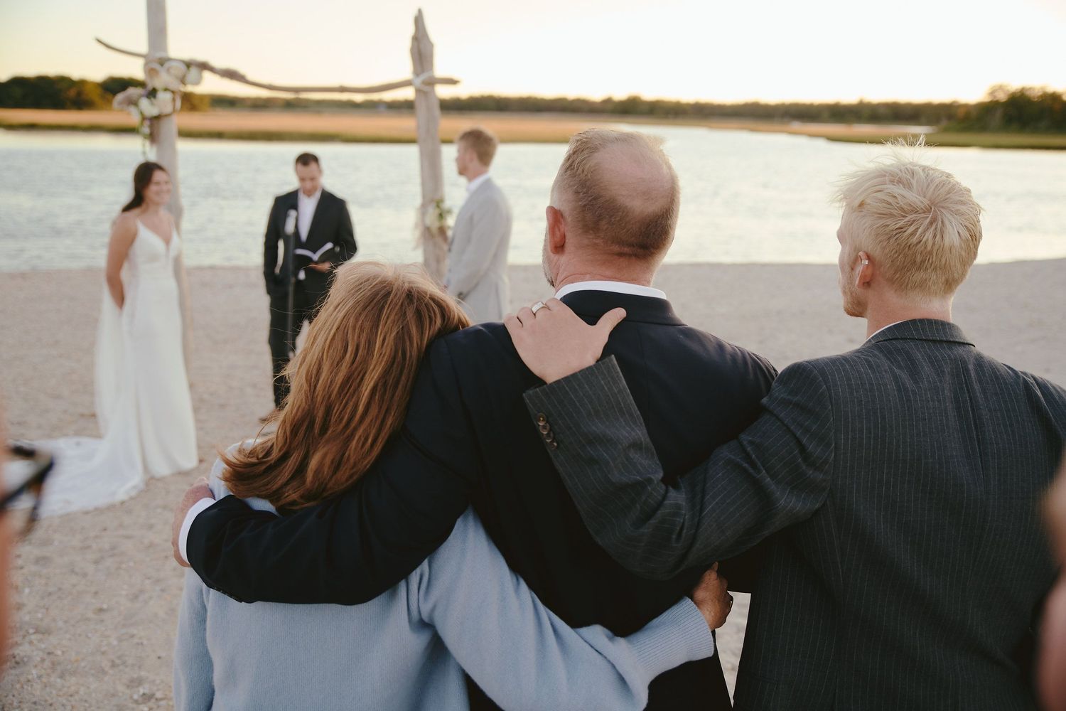 A sunset wedding ceremony takes place by the water as guests watch from behind in a group embrace.
