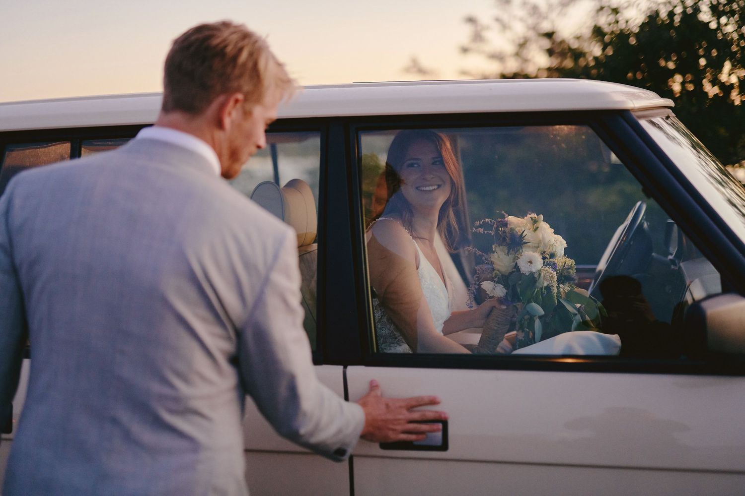 A formal interaction between elegantly dressed guests in a car during sunset at a wedding celebration.