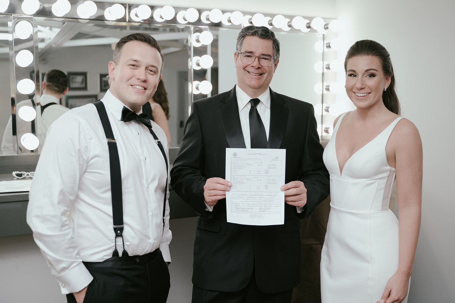 A couple in wedding attire poses with an officiant holding a document in front of a dressing room mirror and lights.