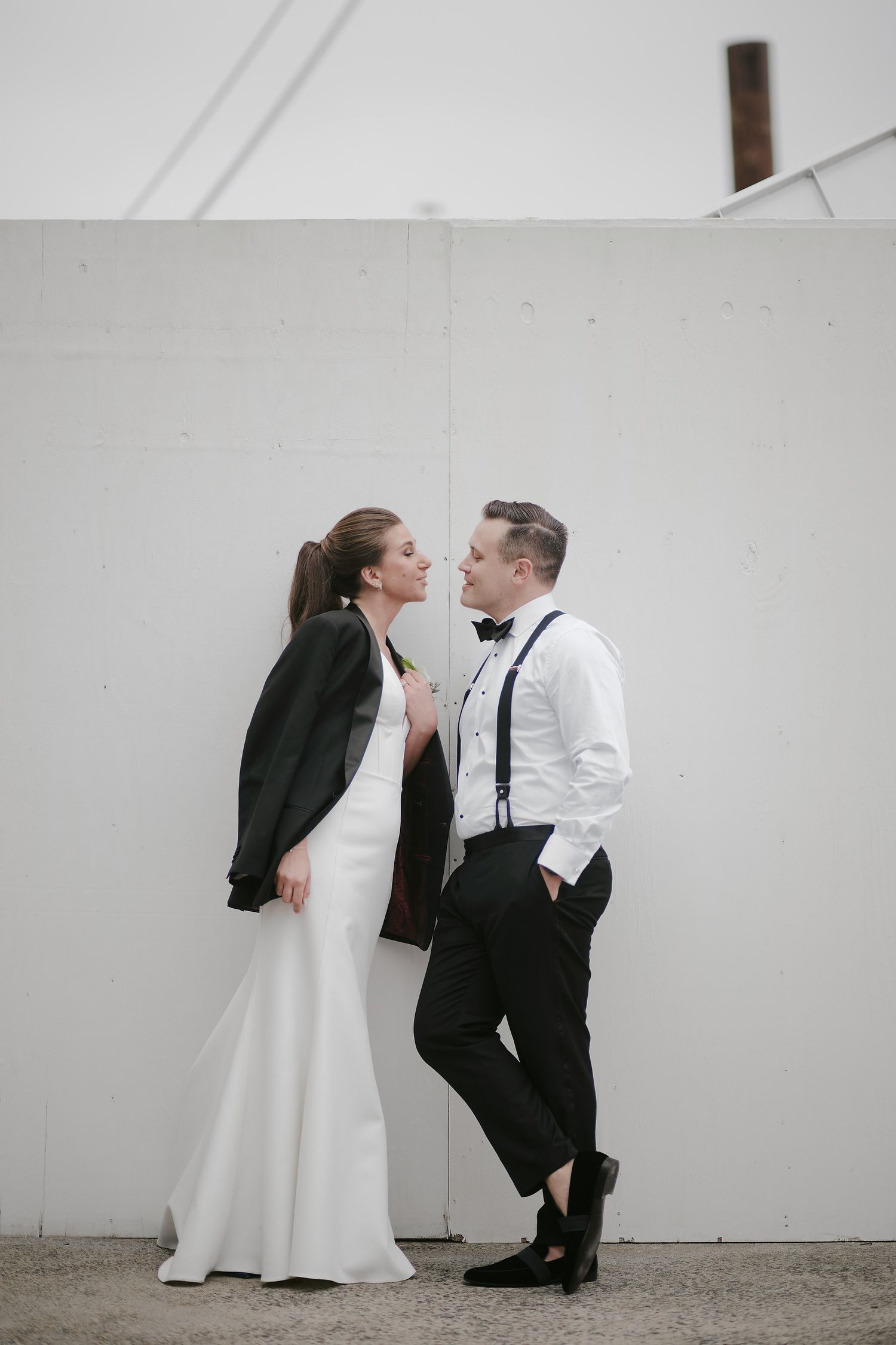 A newlywed couple in elegant black and white attire pose against a white wall in a candid romantic moment.