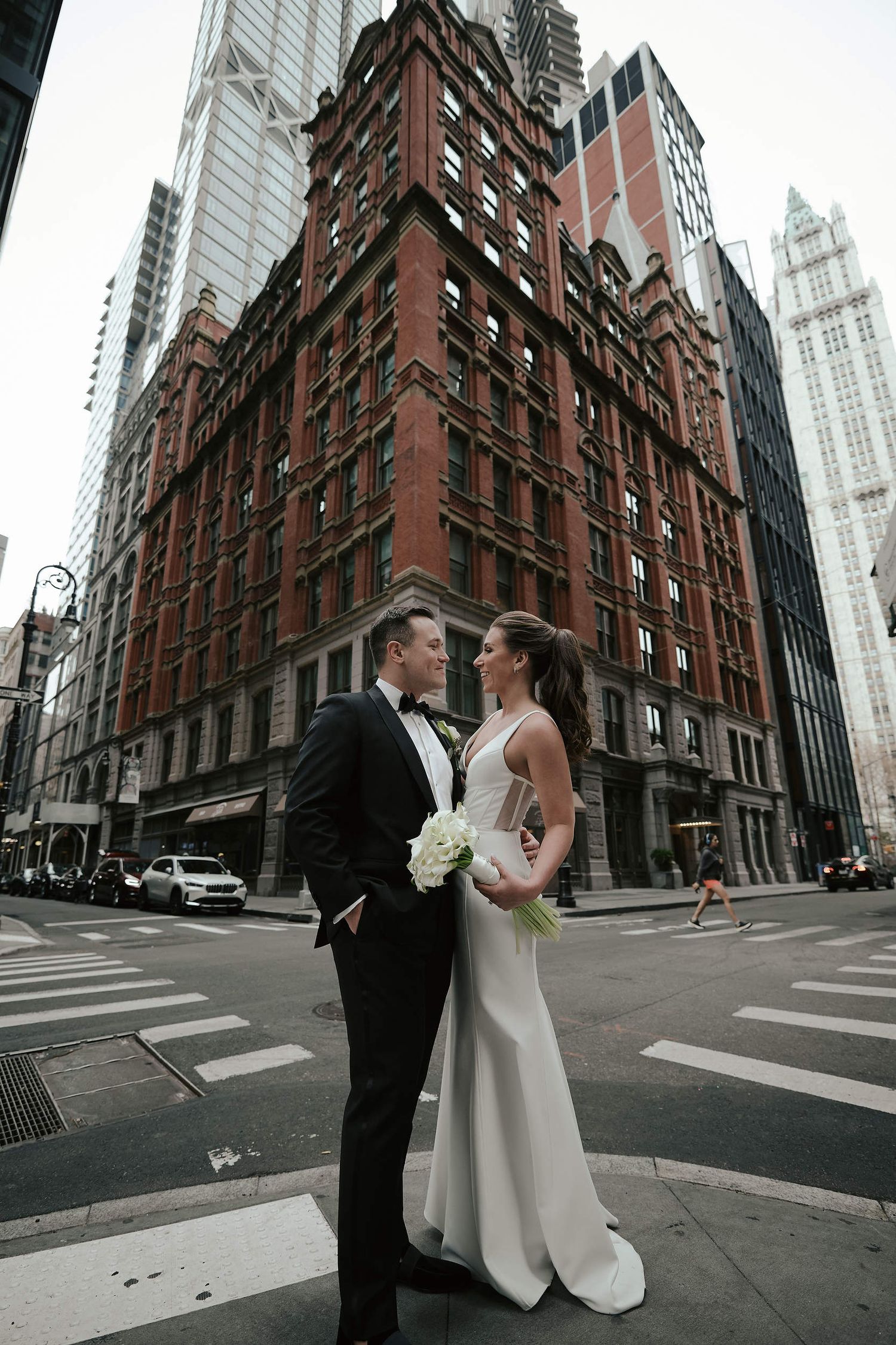 A couple in wedding attire standing on a city street corner with a historic red brick building and skyscrapers in Manhattan.