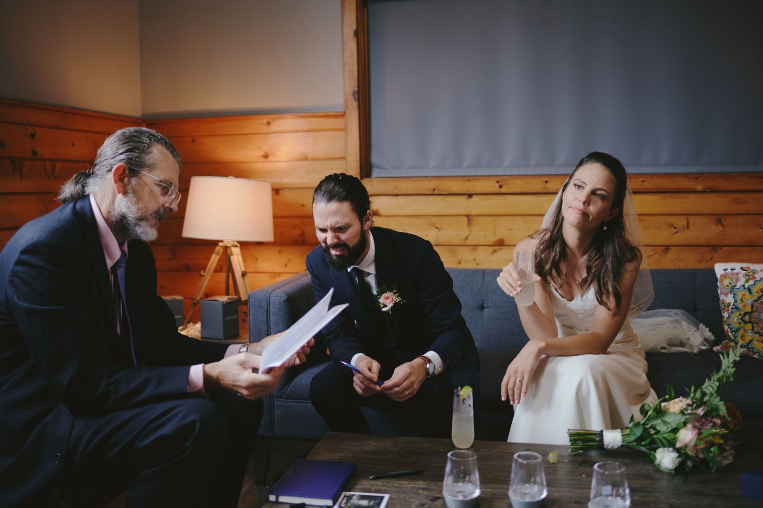 Couple exchanging wedding vows indoors with officiant reading from papers in a cozy wood-paneled room.