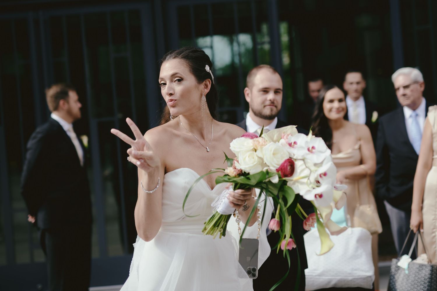 A bride in a white gown flashes a peace sign while holding a pink and white bouquet at a wedding celebration.
