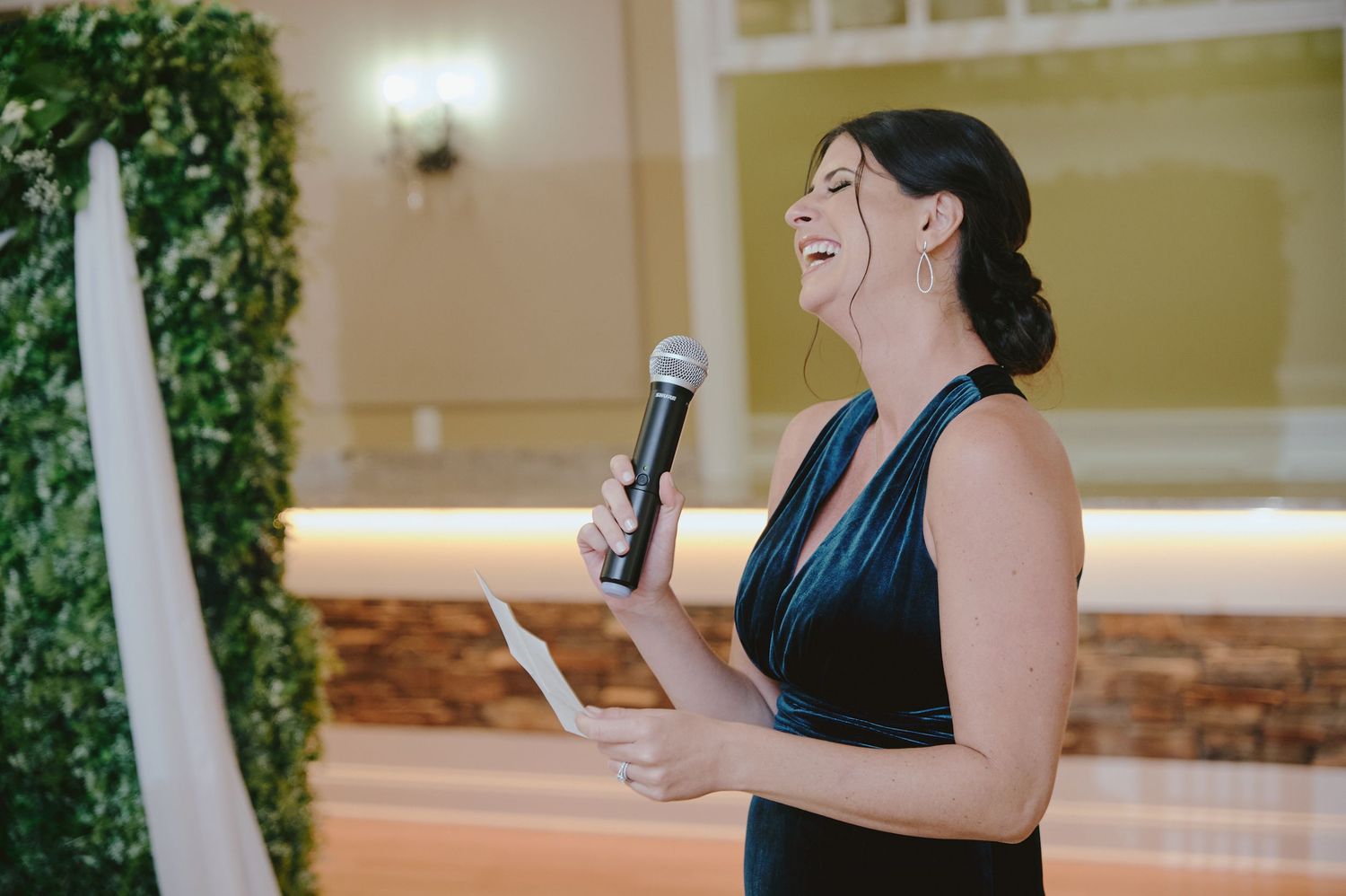 A person in a navy dress giving a speech with a microphone while holding papers at an elegant indoor venue.