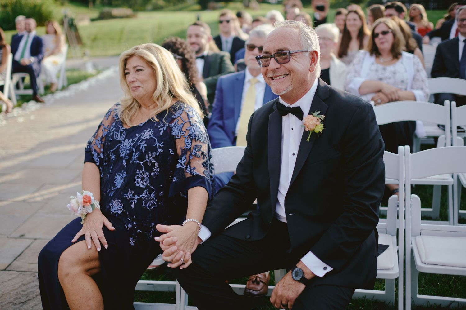Guests share a joyful moment while seated at an outdoor wedding ceremony with white chairs and greenery in background.