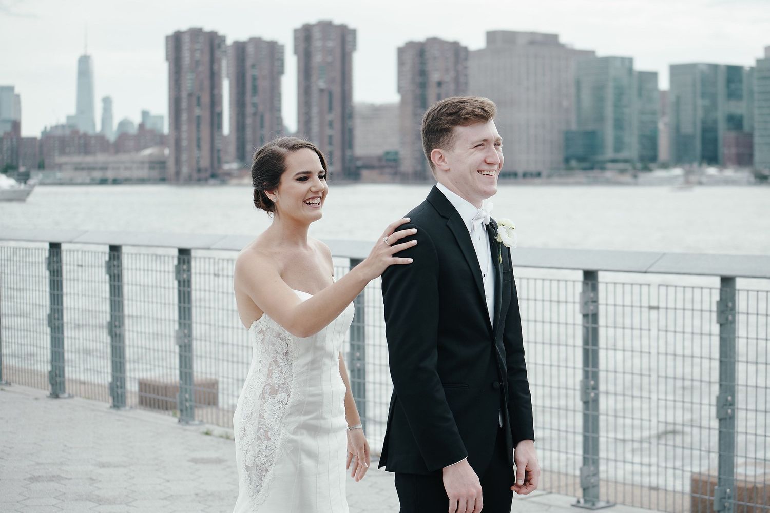 A bride and groom pose along a waterfront railing with the New York City skyline in the background.