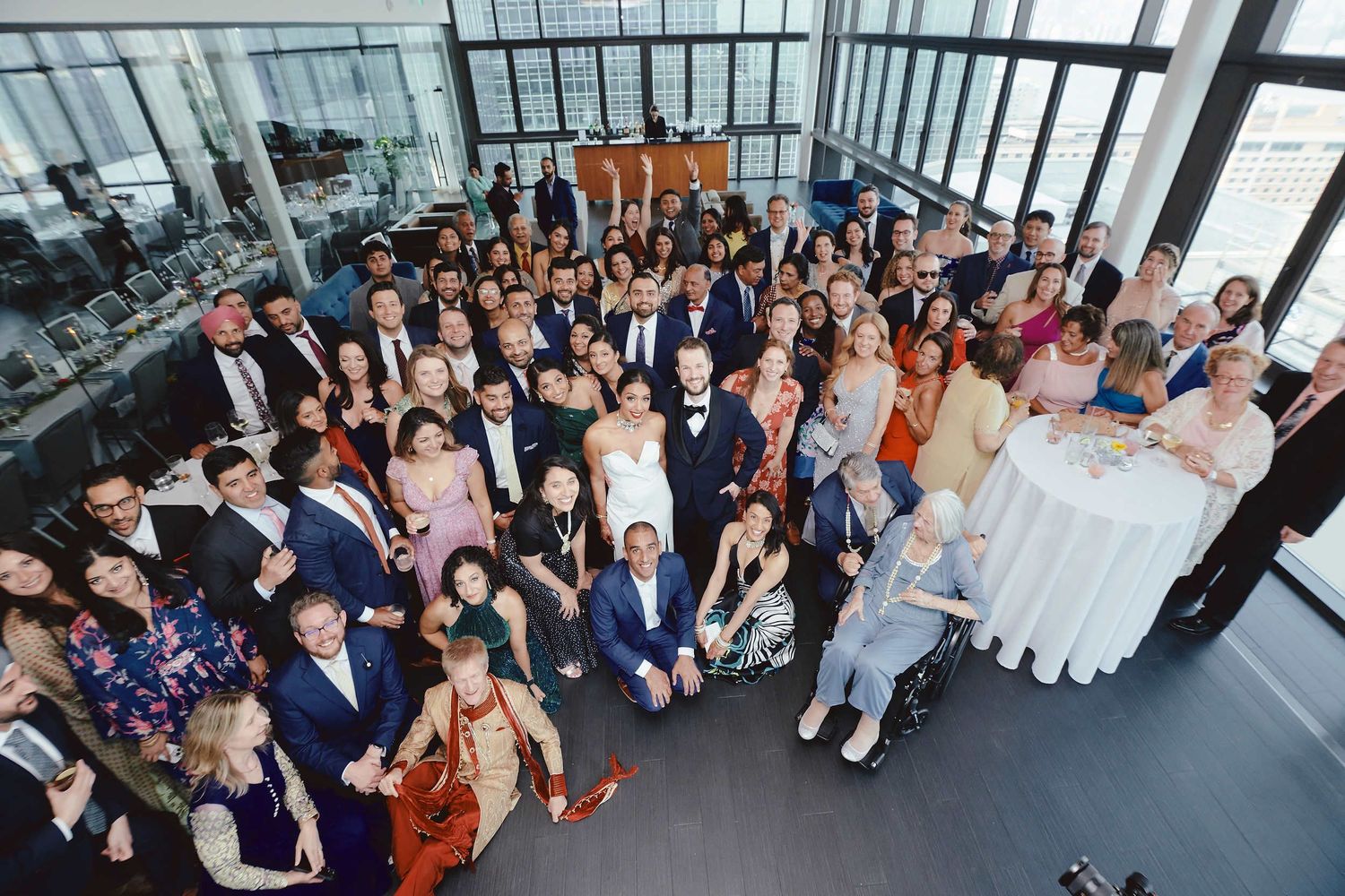 Large group photo of wedding guests and attendees posing together in a bright modern venue with city views.