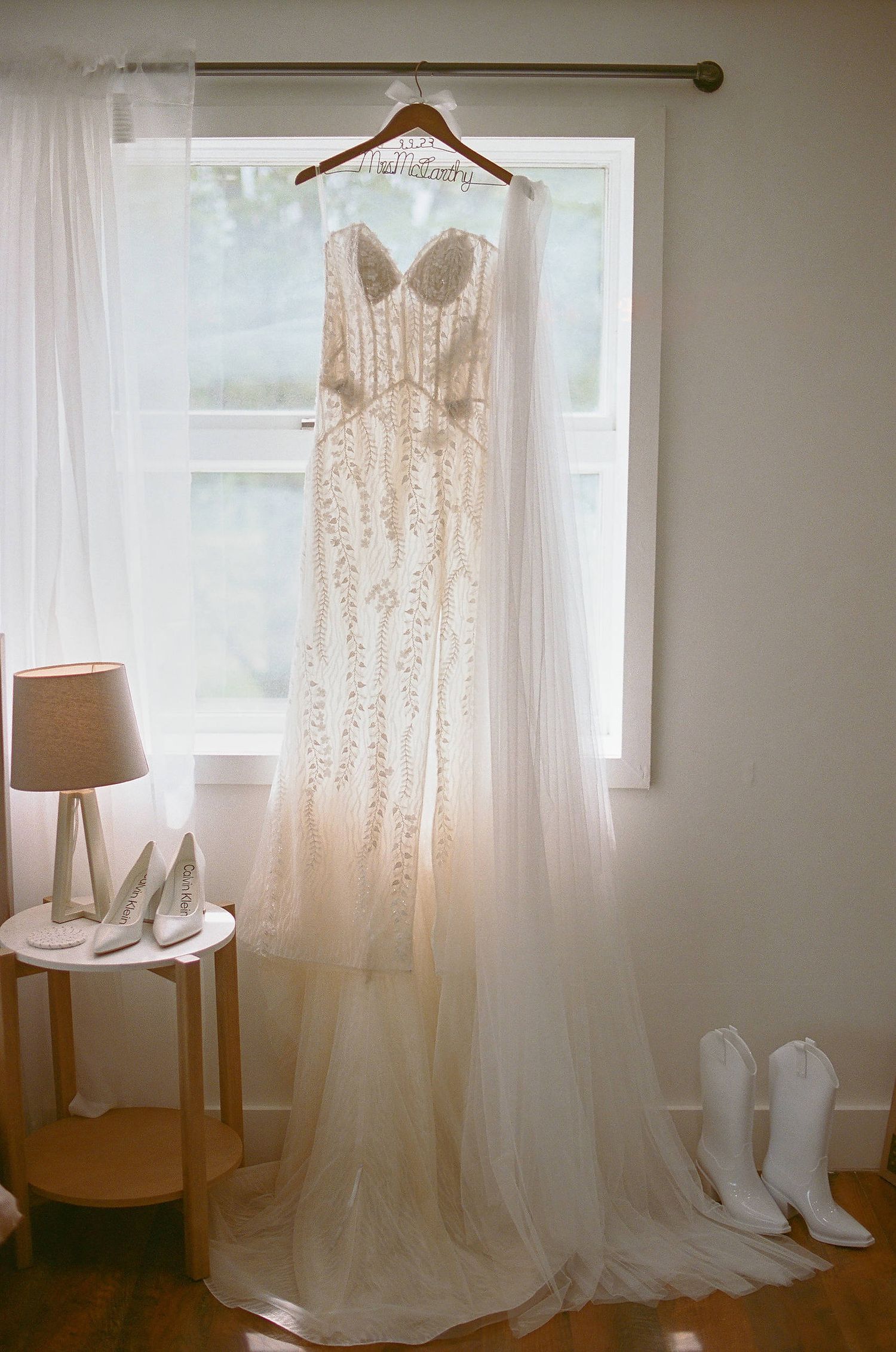 Elegant white lace wedding dress hangs by window with bridal boots and table lamp creating romantic getting ready scene.
