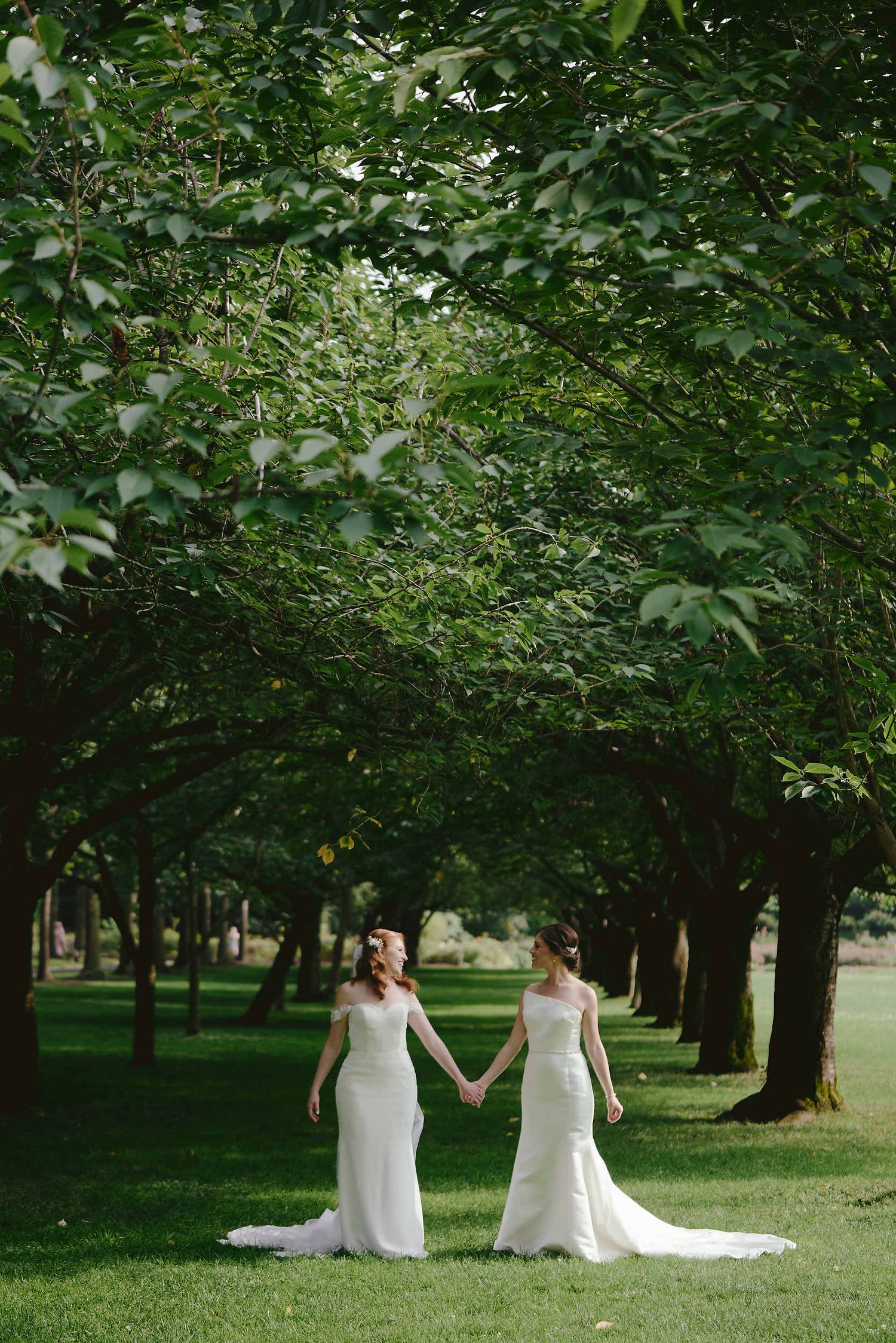 Two people in elegant white wedding gowns hold hands while standing in a tree-lined garden path on a sunny day.