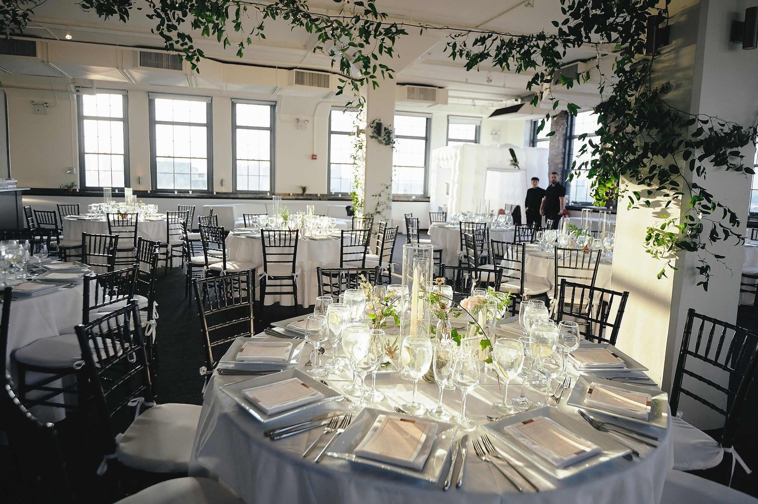 Elegant wedding reception venue with round tables, chiavari chairs, and white linens in a bright room with large windows.