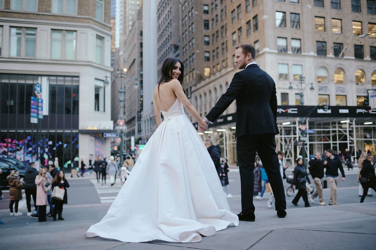 A couple in wedding attire stand together on a busy city sidewalk with tall buildings in the background.