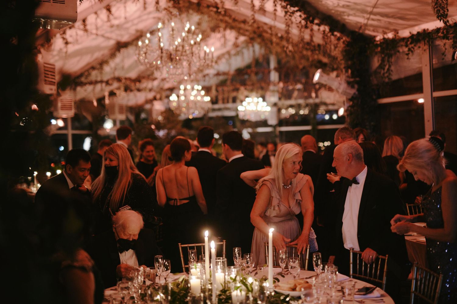 A dimly lit elegant reception venue with crystal chandeliers and candlelit tables as guests mingle and dine at night.