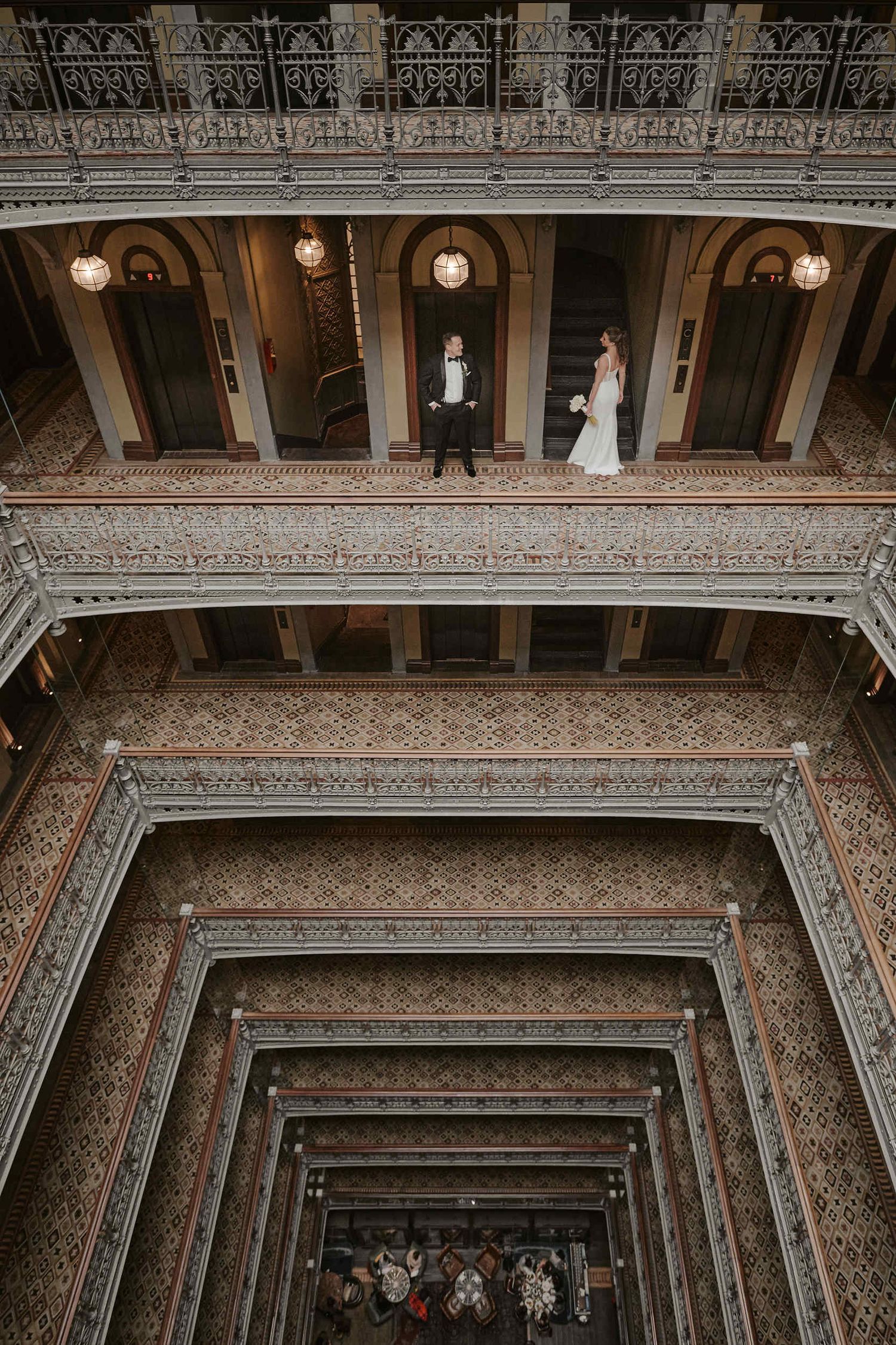 Wedding couple stands on ornate tiered balconies of historic Beekman Hotel while guests gather below in atrium.