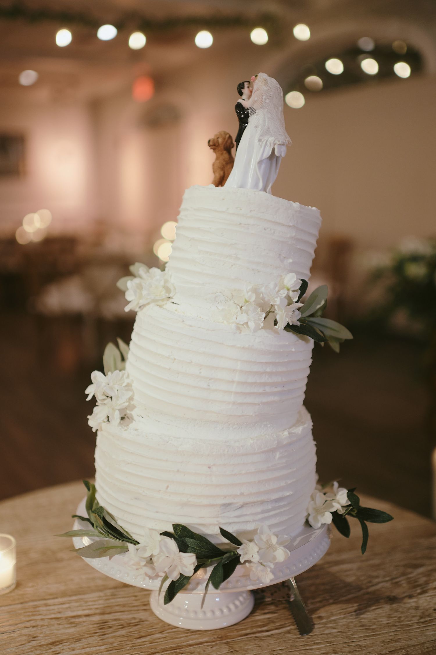 Three-tier white buttercream wedding cake decorated with fresh white flowers and greenery on a rustic wooden table.