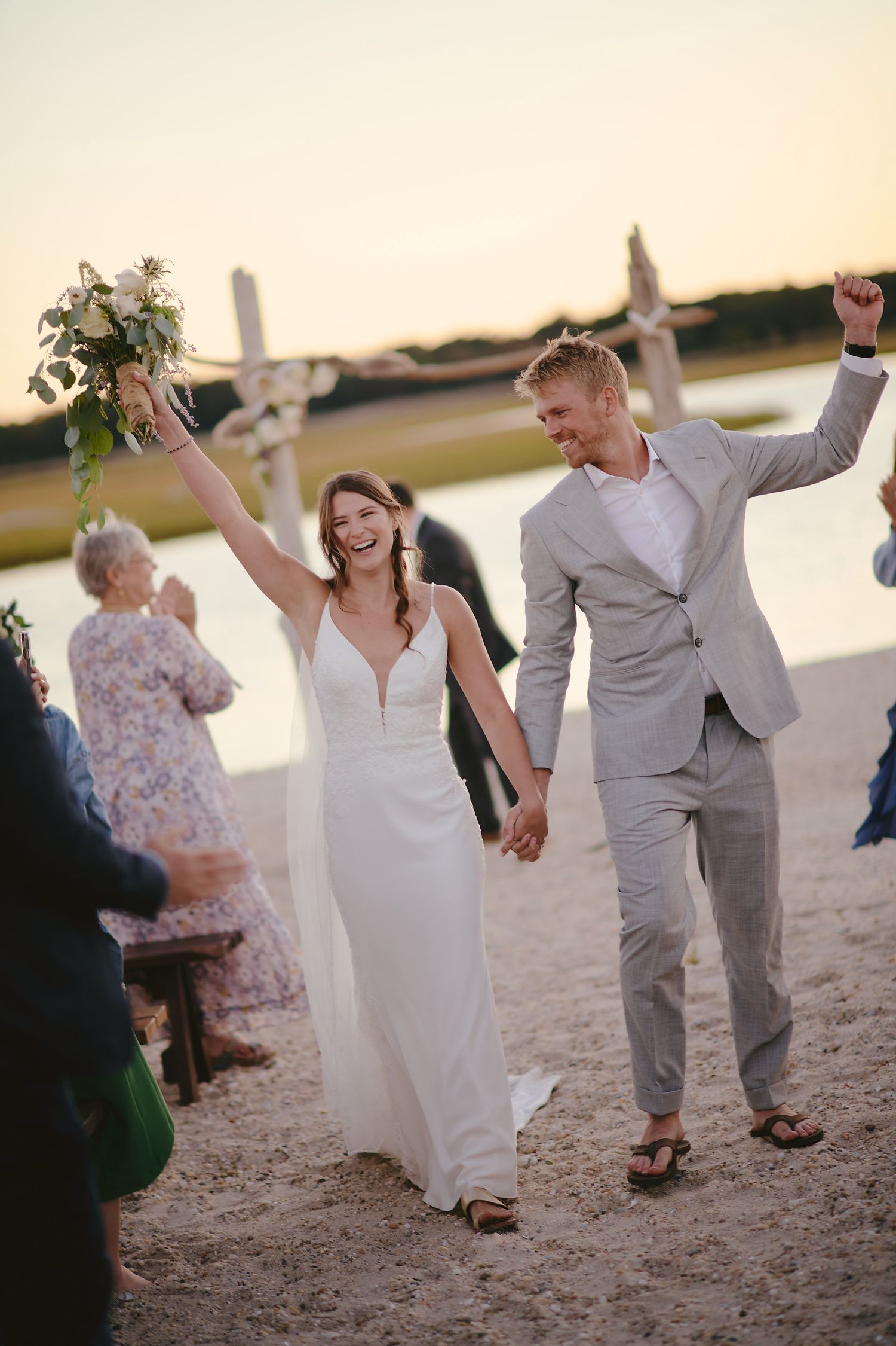 Newlyweds celebrate joyfully while walking on a beach at sunset after their wedding ceremony.
