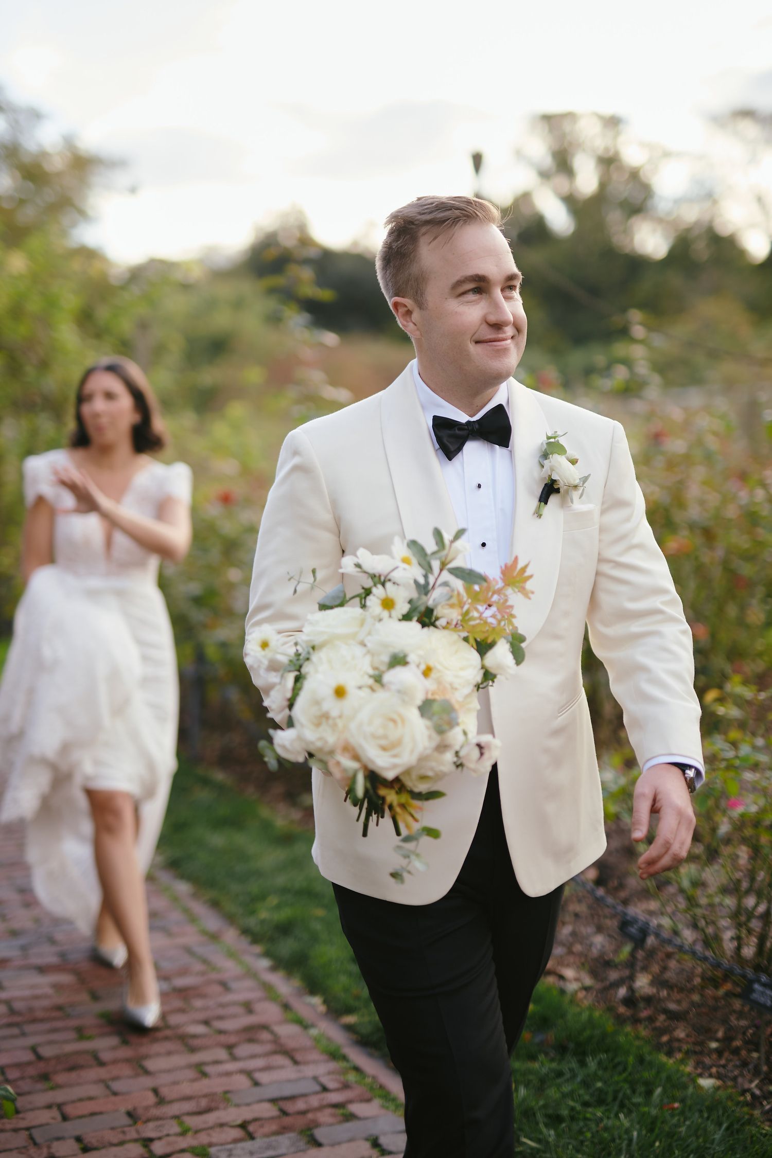 A person in a white tuxedo jacket carries a beautiful bouquet of white roses along a brick pathway.