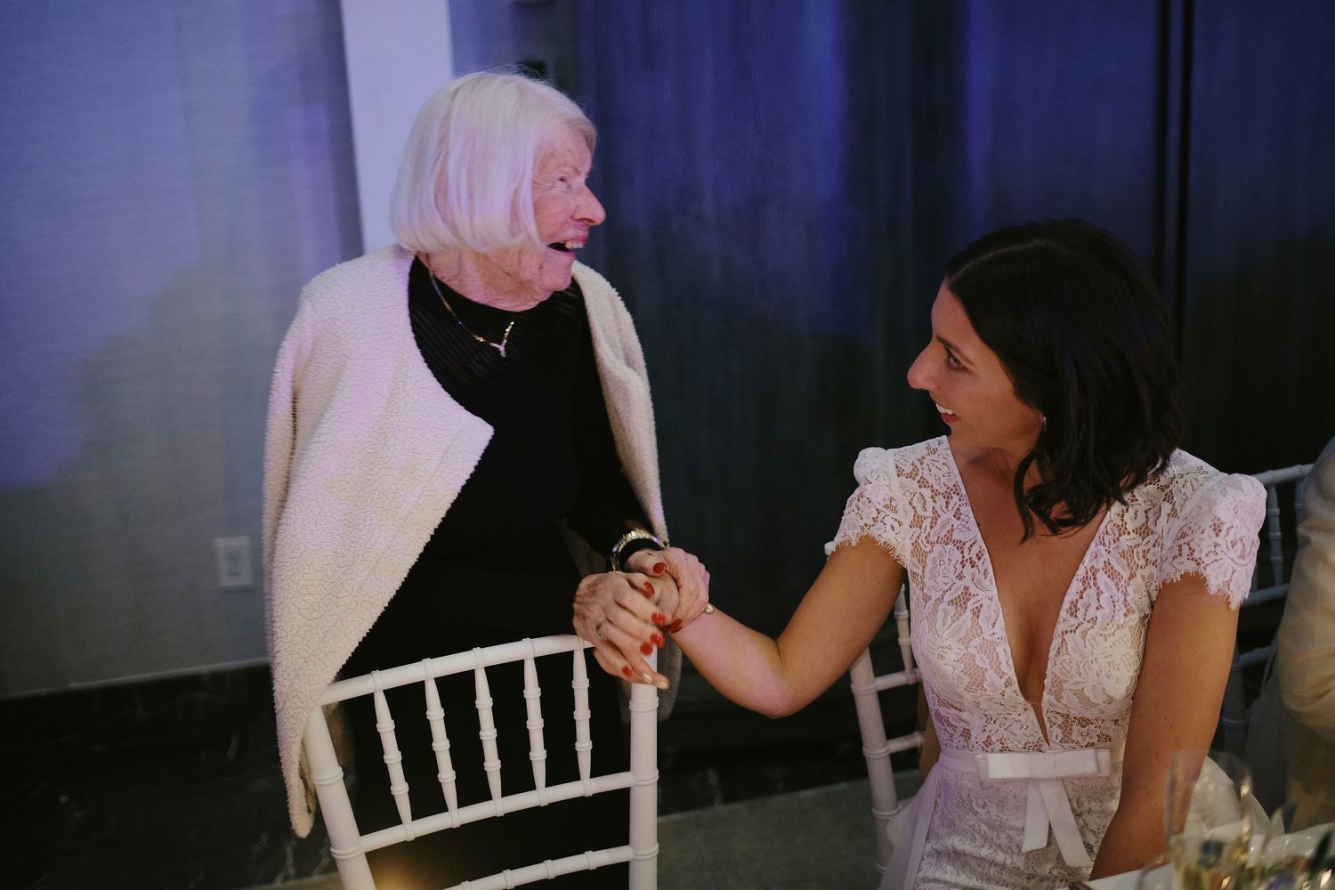 Two people share an emotional moment, holding hands while seated at an event space with white chairs.
