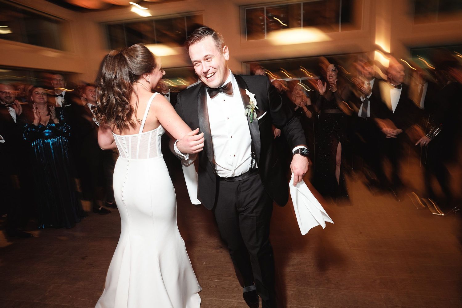 A couple dances together at their wedding reception in formal attire as guests look on in the background.