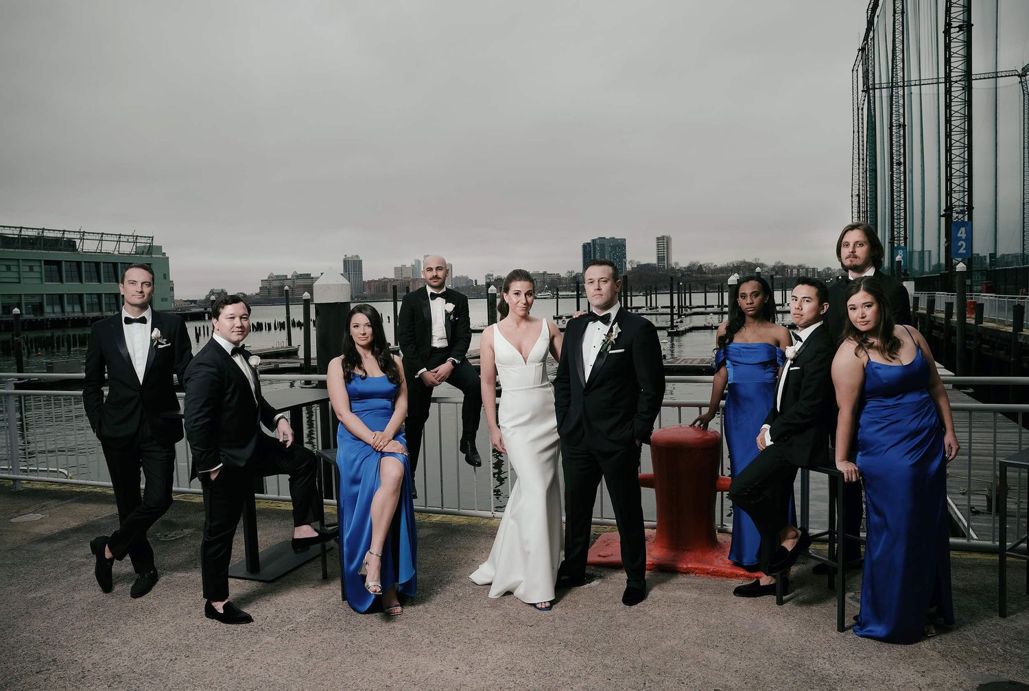 A bridal party in formal blue dresses and black tuxedos poses on a rooftop terrace with city skyline views.