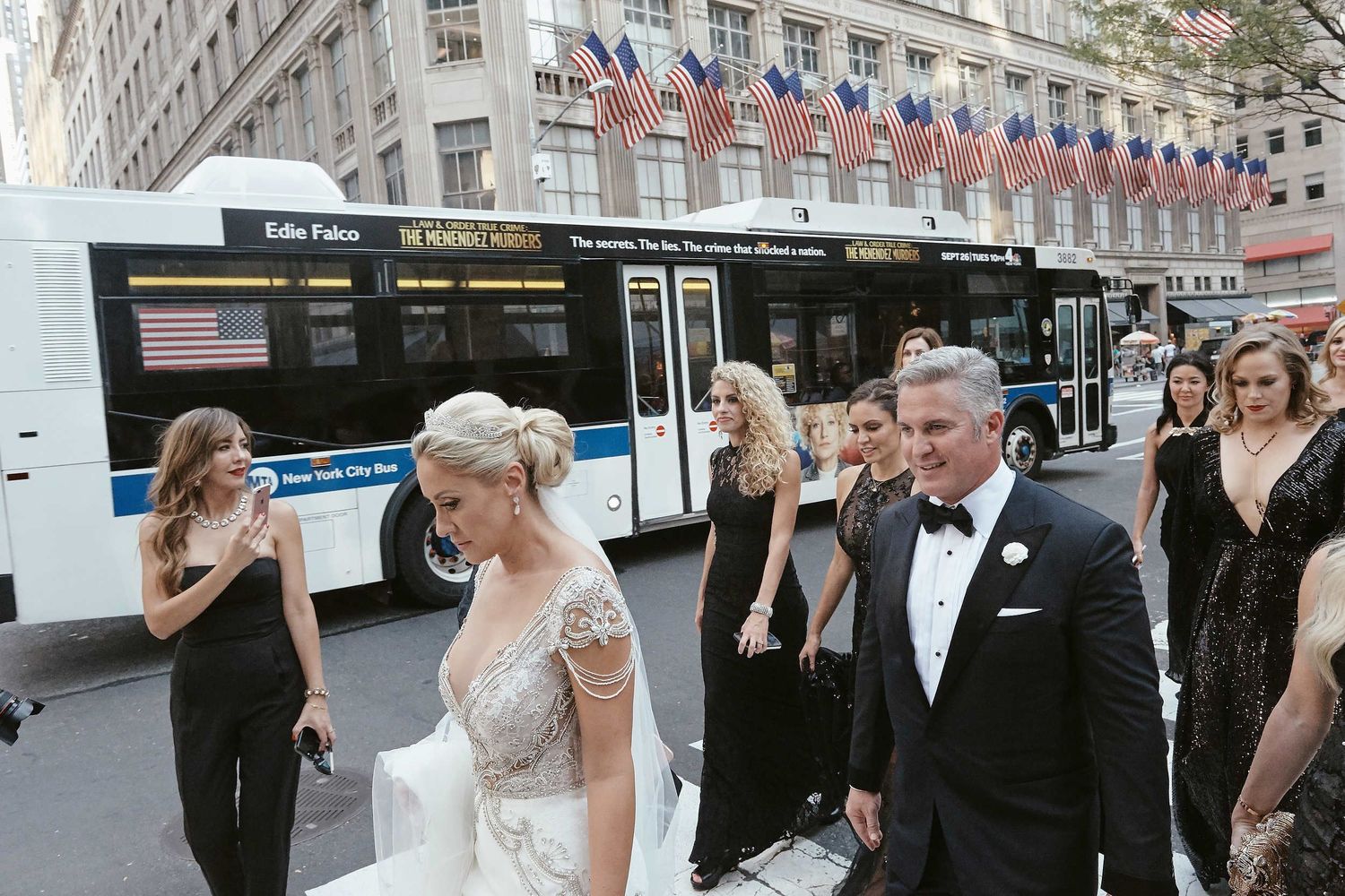 Wedding party in formal attire walking near a city bus with American flags hanging on the building in the background.