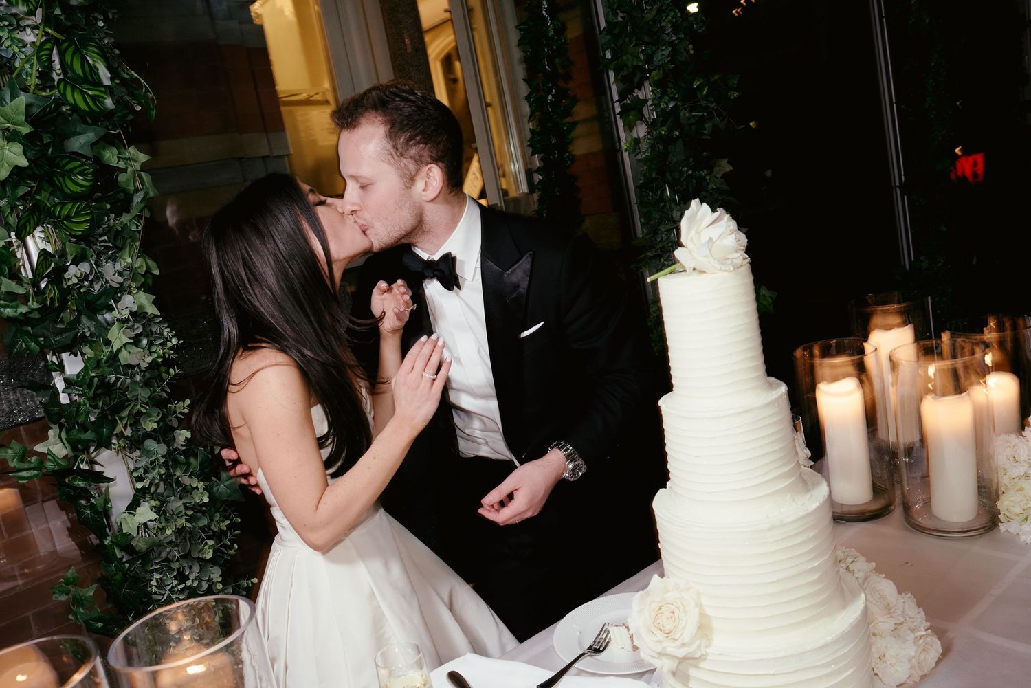 Elegant wedding couple shares a kiss next to their white tiered wedding cake adorned with candles and greenery.