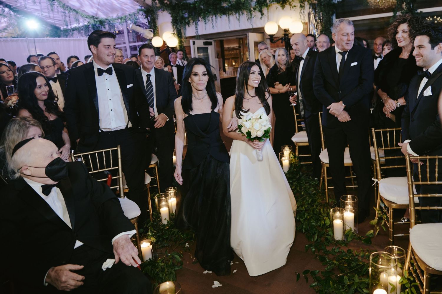 Indoor wedding ceremony with bride walking down candlelit aisle among guests in formal attire at evening event.