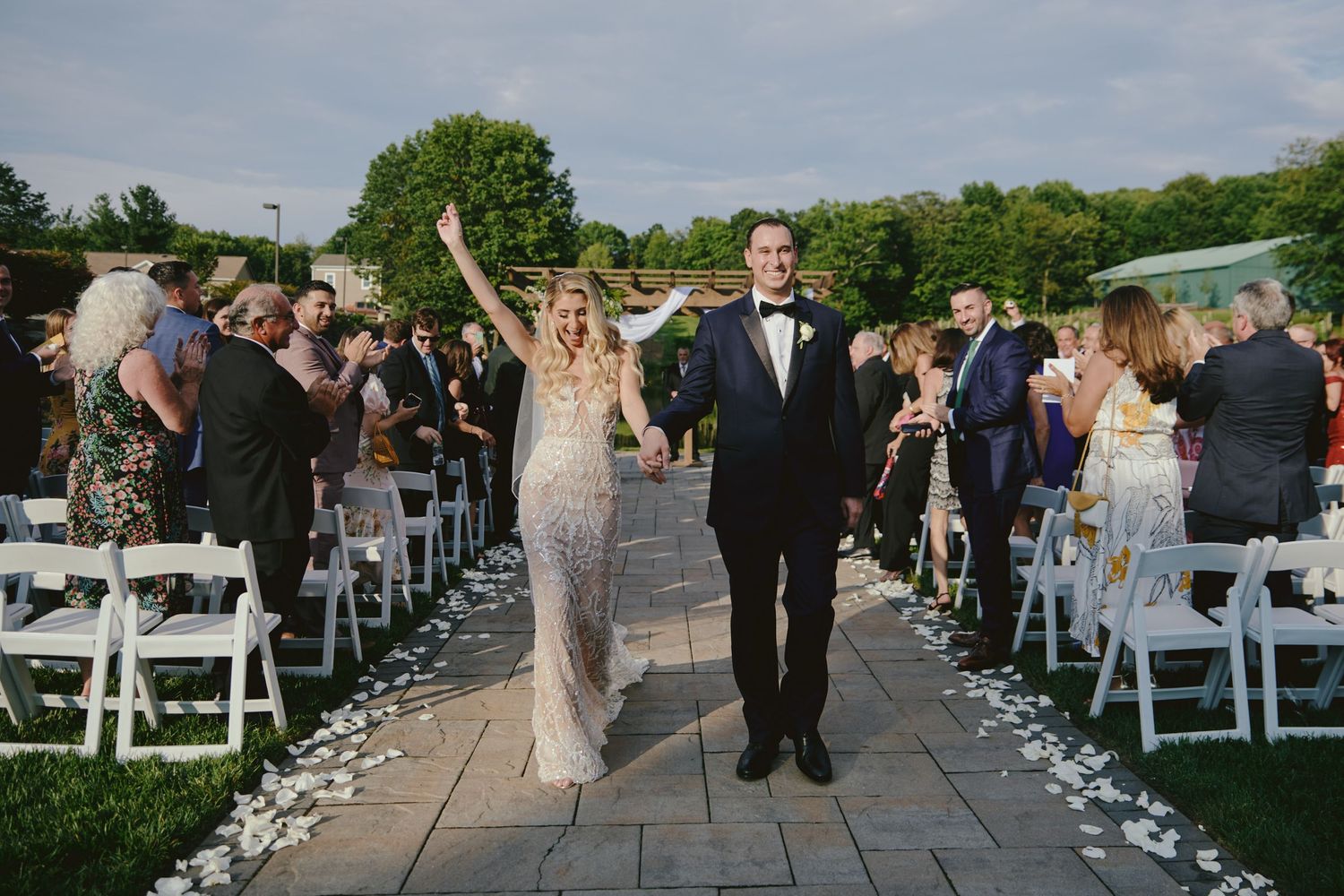 A joyous celebration as a newlywed couple walks down an outdoor aisle lined with flower petals and white chairs.
