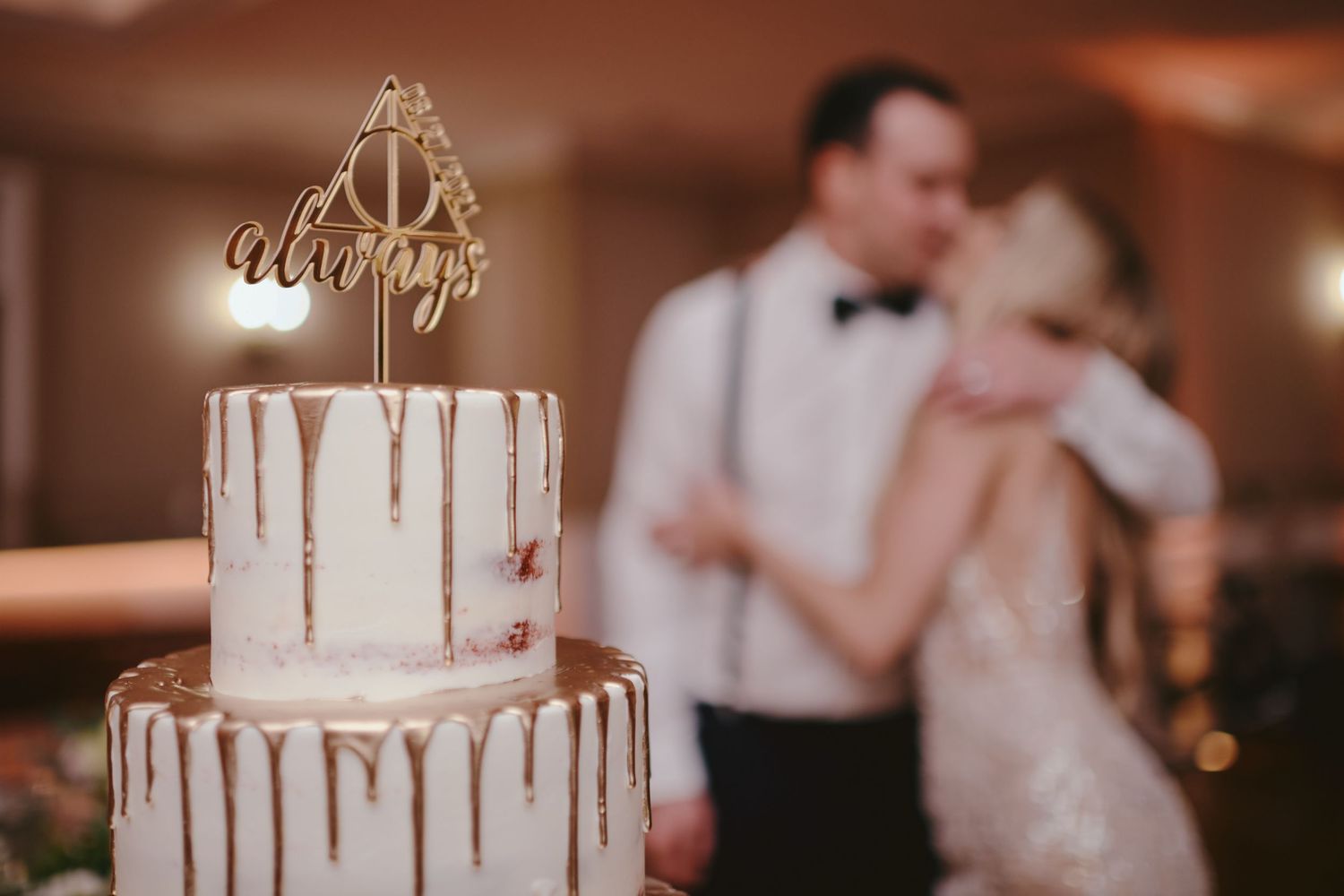 Elegant white wedding cake with gold drip frosting sits in focus while couple dances blurred in background.
