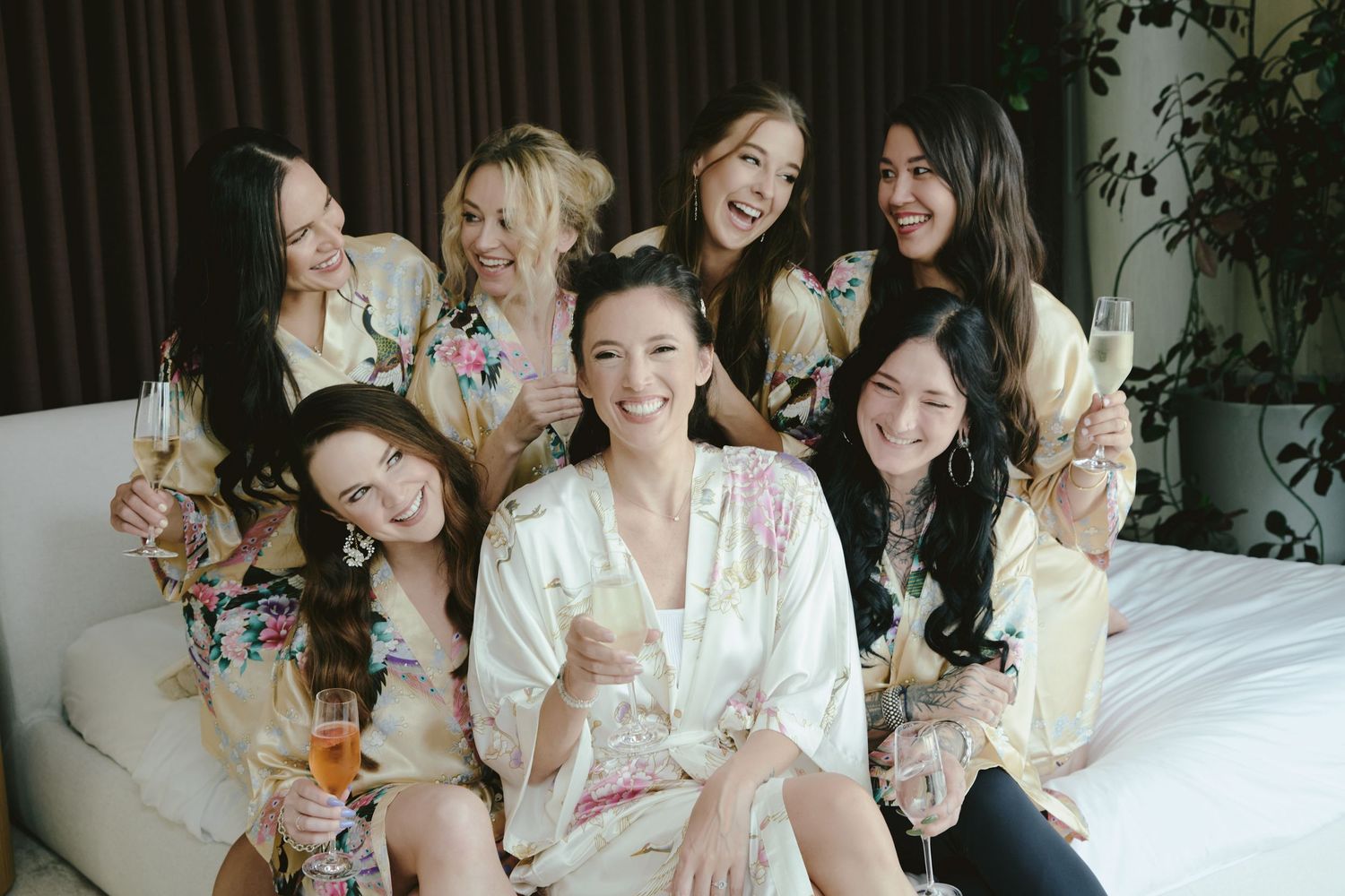 A group of friends in silky robes enjoy champagne while sitting together on a white couch at a special celebration.
