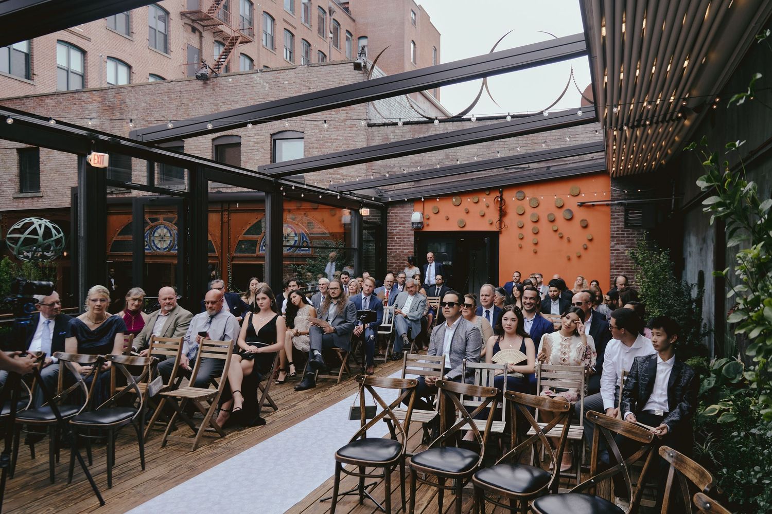 Wedding guests seated in rustic wooden chairs at an outdoor urban venue with industrial pergola and string lights overhead.
